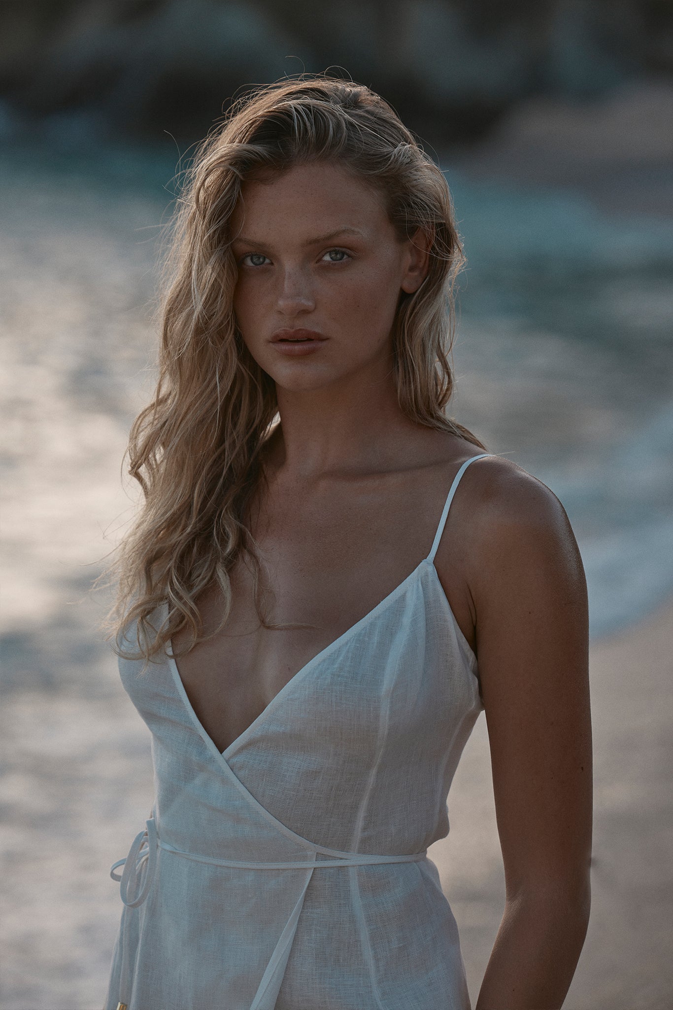 Young woman on a sandy beach at dusk, wavy blonde hair, white linen wrap dress with thin straps, neutral expression