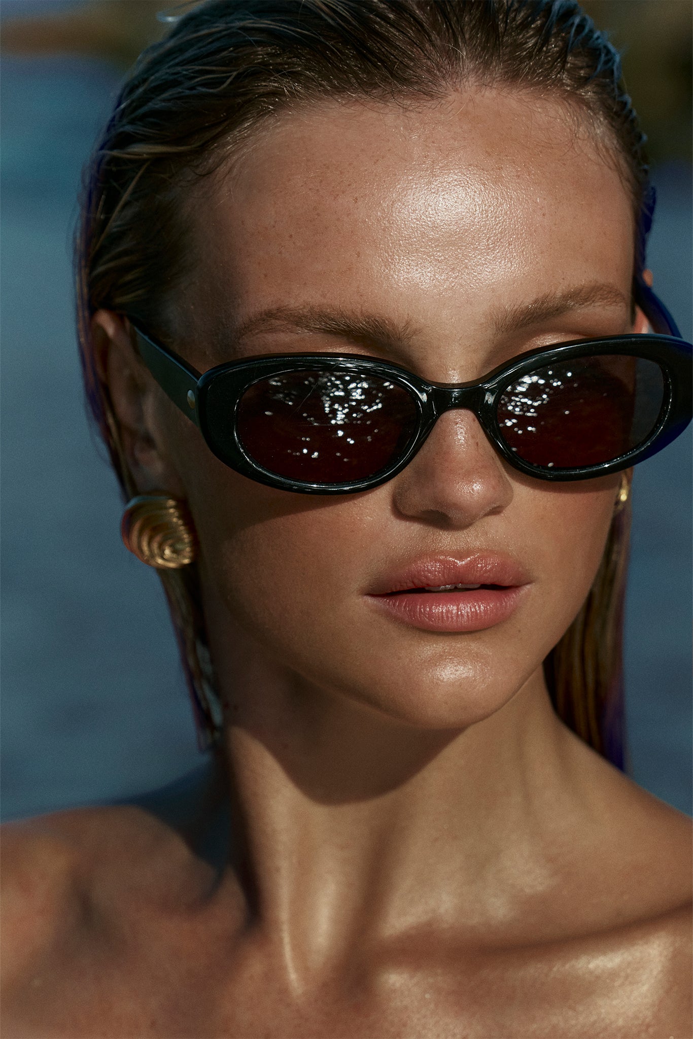 Close-up portrait of a woman with wet hair, glossy skin, black oval sunglasses reflecting light, and a gold spiral earring.