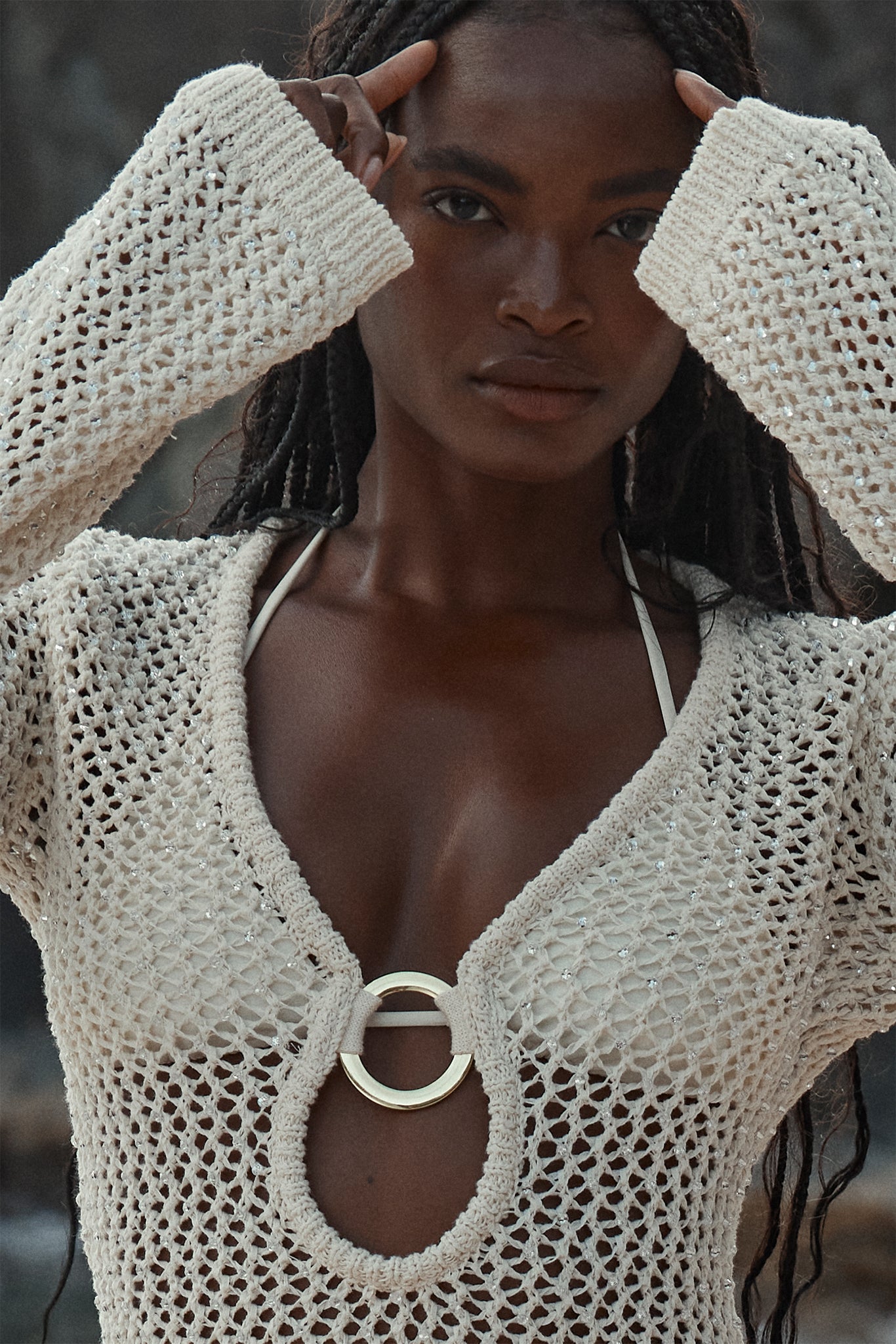 Woman wearing cream open-knit crochet top with circular metal ring at chest over white bikini straps, hands near forehead
