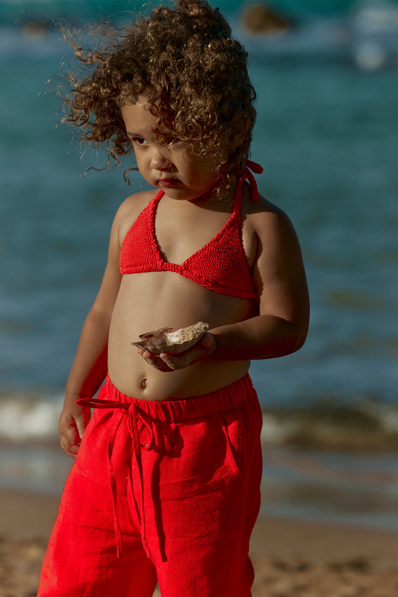 Young child with curly hair wearing red bikini top and red pants, holding a seashell at the shoreline.