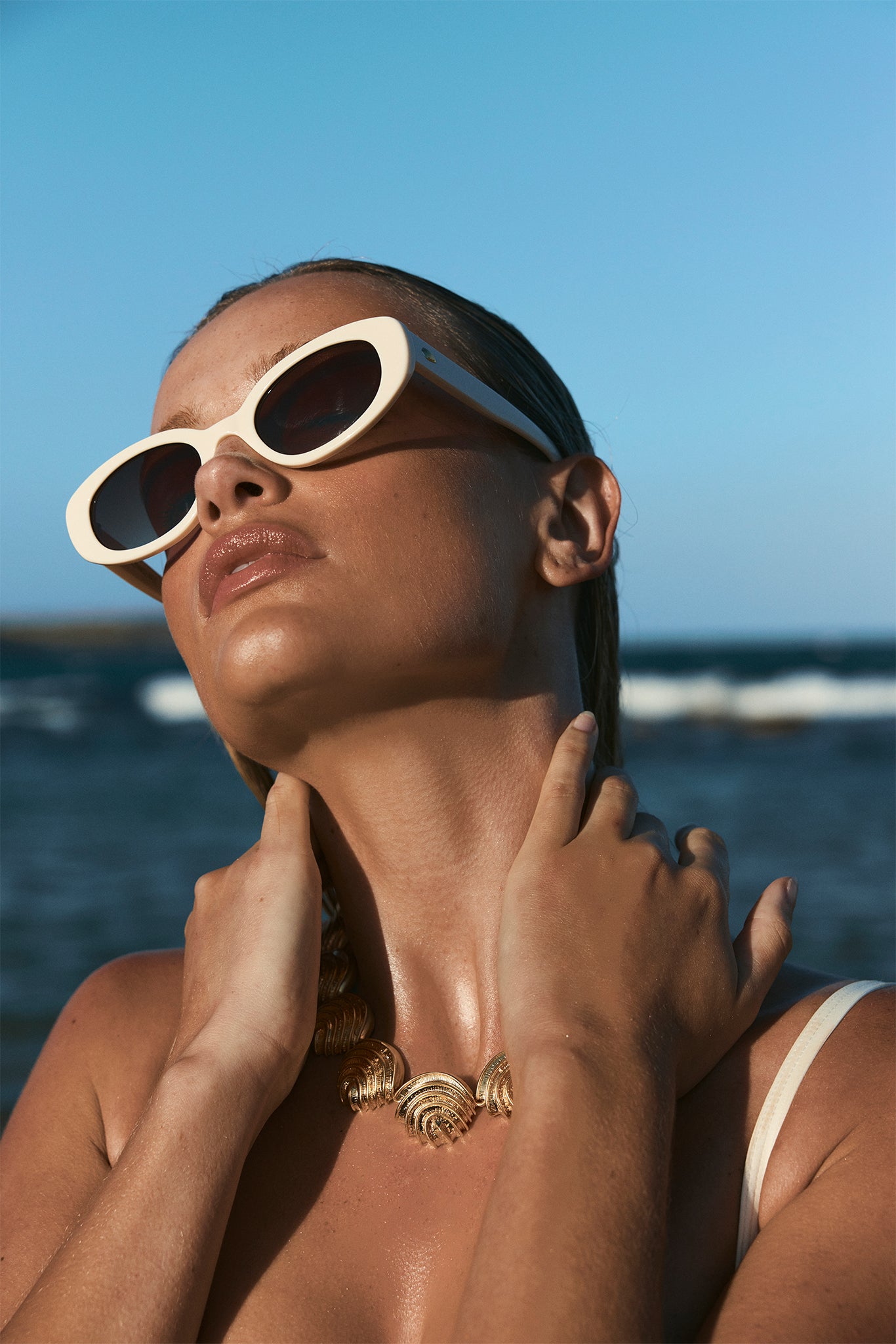 Woman wearing white cat-eye sunglasses and a gold shell necklace at the beach, head tilted back toward sky.