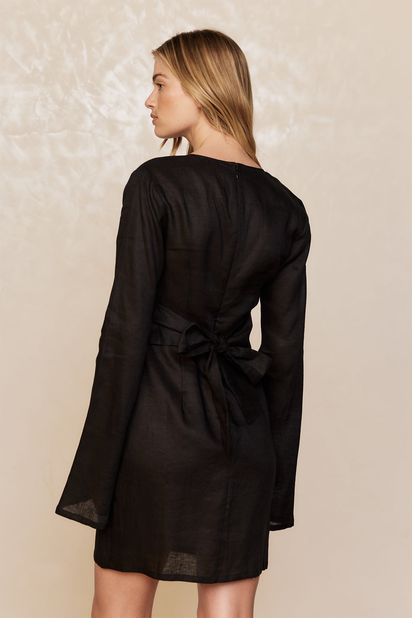 Woman shown from the back wearing a black long-sleeve linen mini dress with a tied waist bow on a neutral background.