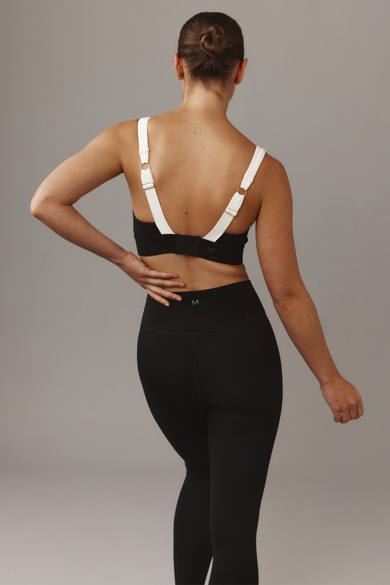 Woman seen from back wearing black sports bra with white straps and high-waist leggings; M logo visible on band.