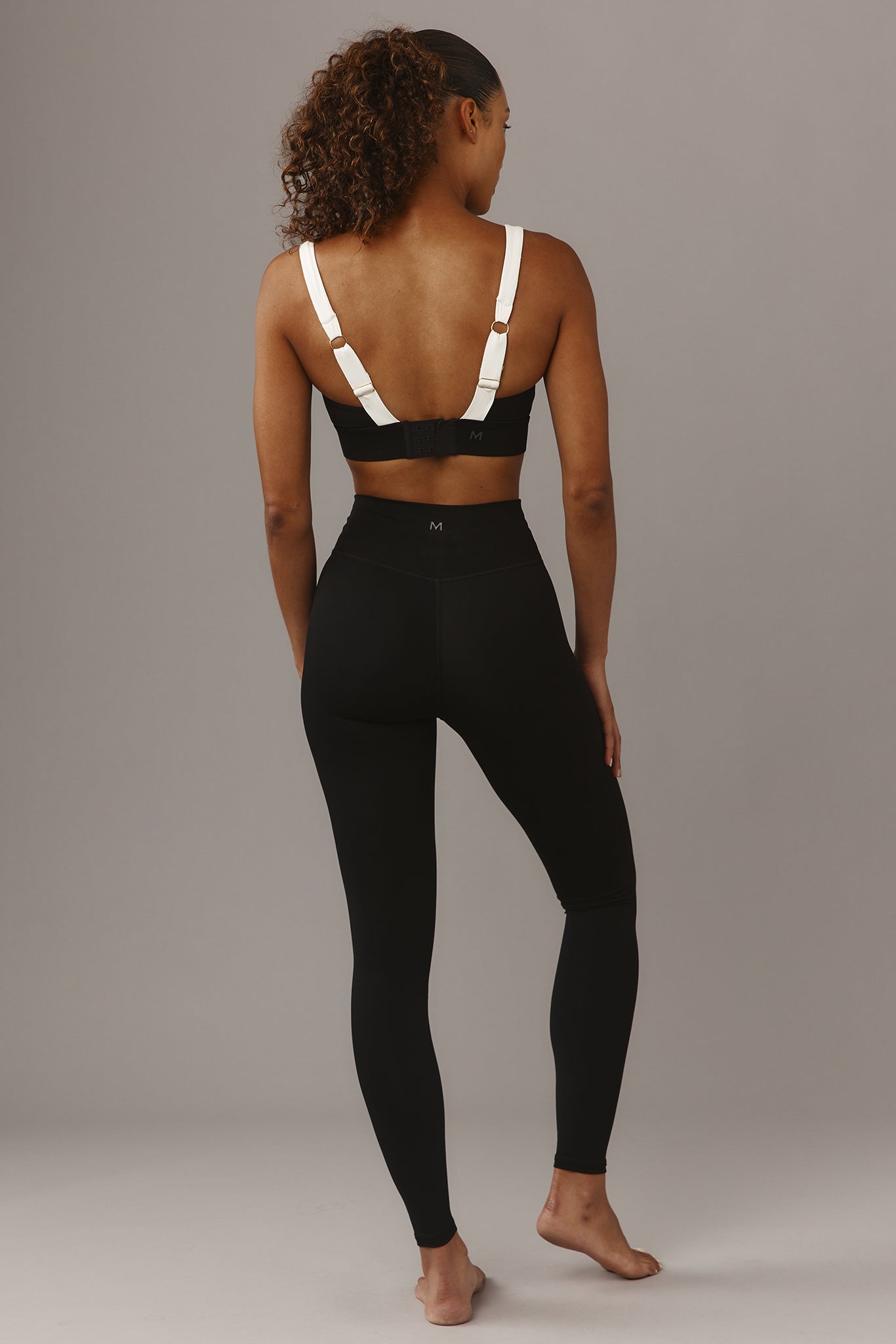 Back view of model wearing black high-waist leggings and black sports bra with white straps; small M logos on bra and waistband.