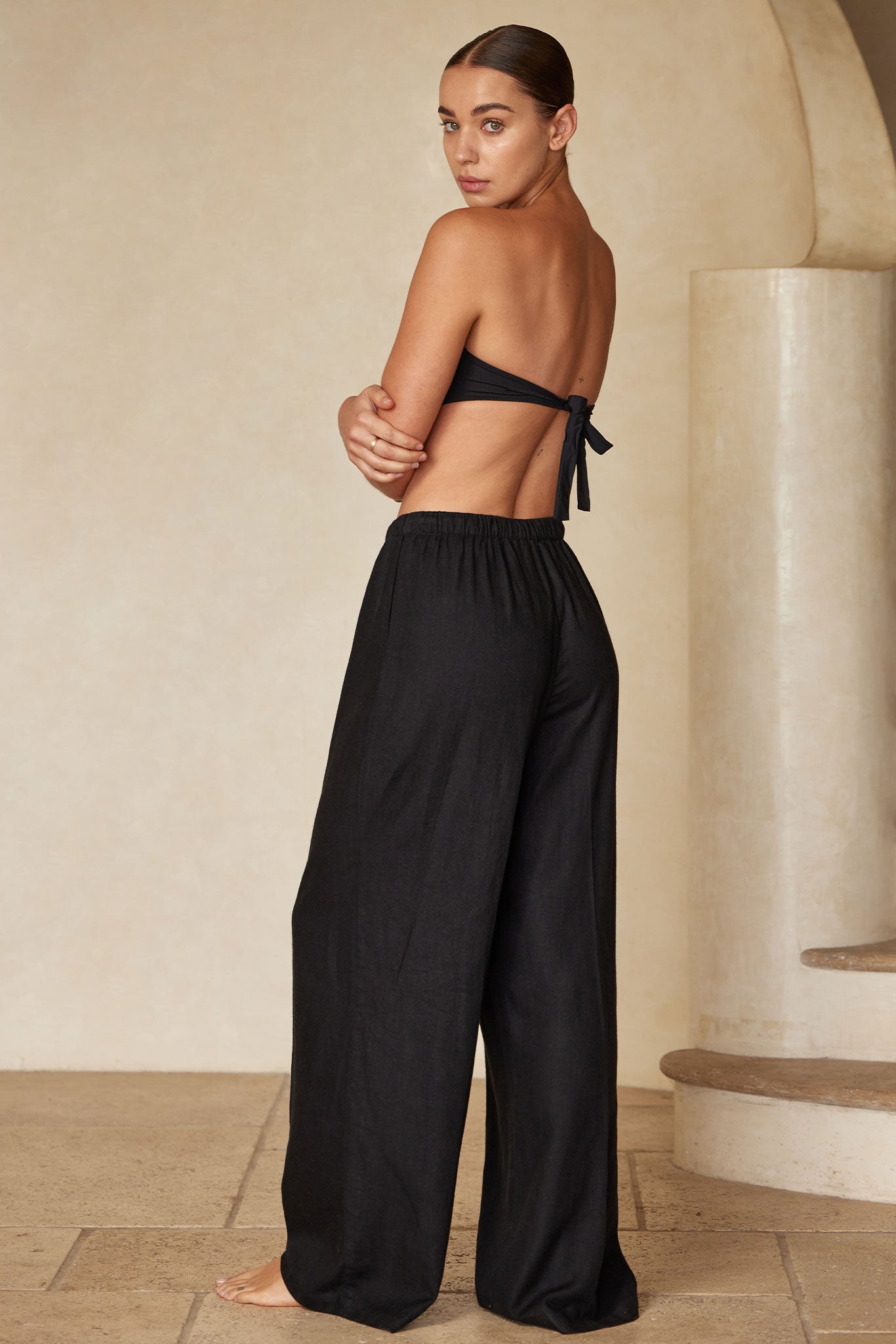 Model back view wearing tied black bandeau top and high-waisted wide-leg black pants, barefoot, looking over her shoulder.