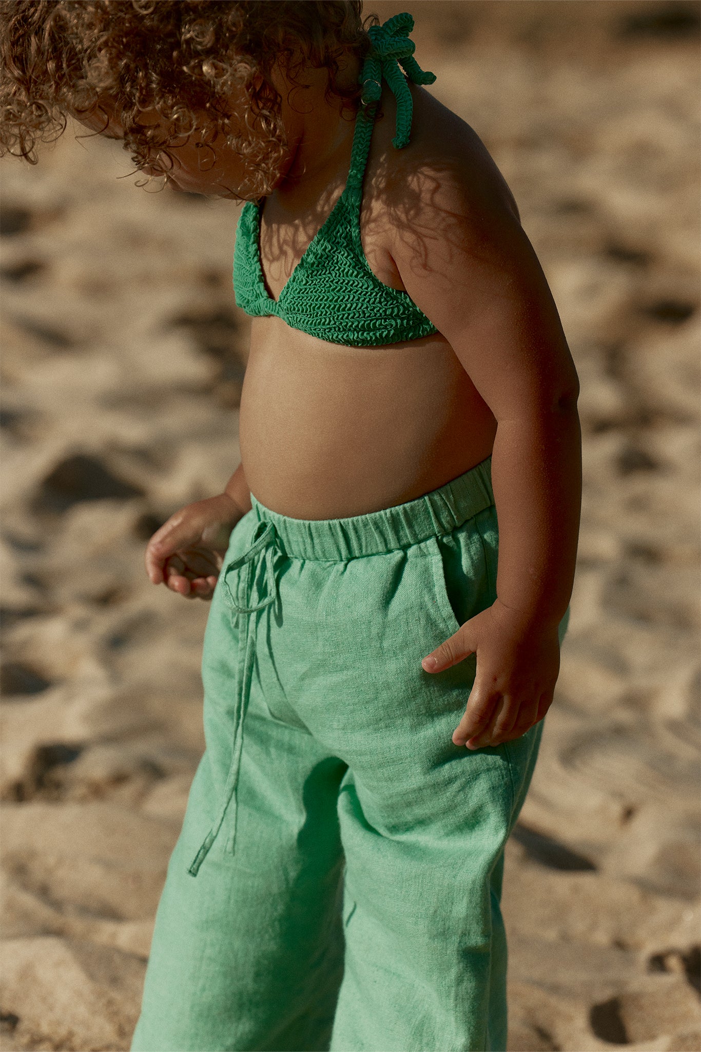 Toddler standing on a sunlit sandy beach wearing a green knitted bikini top and loose mint-green drawstring pants.
