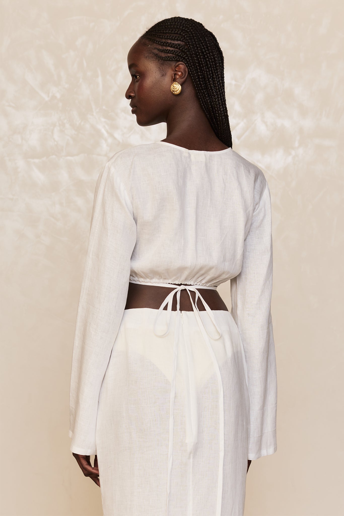 Rear view of woman with long braids wearing a white linen tie-back crop top and matching skirt, gold button earrings.
