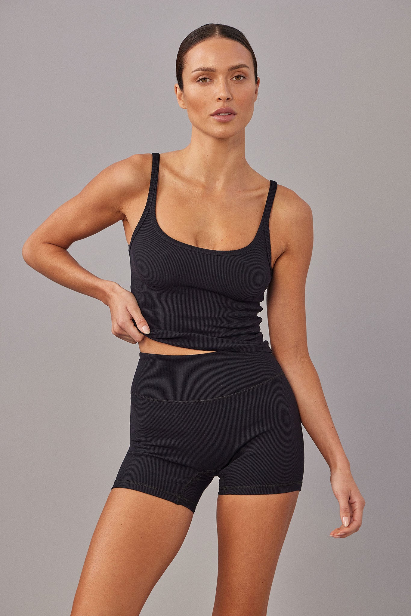Woman modeling black ribbed tank top and high-waist ribbed shorts, tugging hem slightly