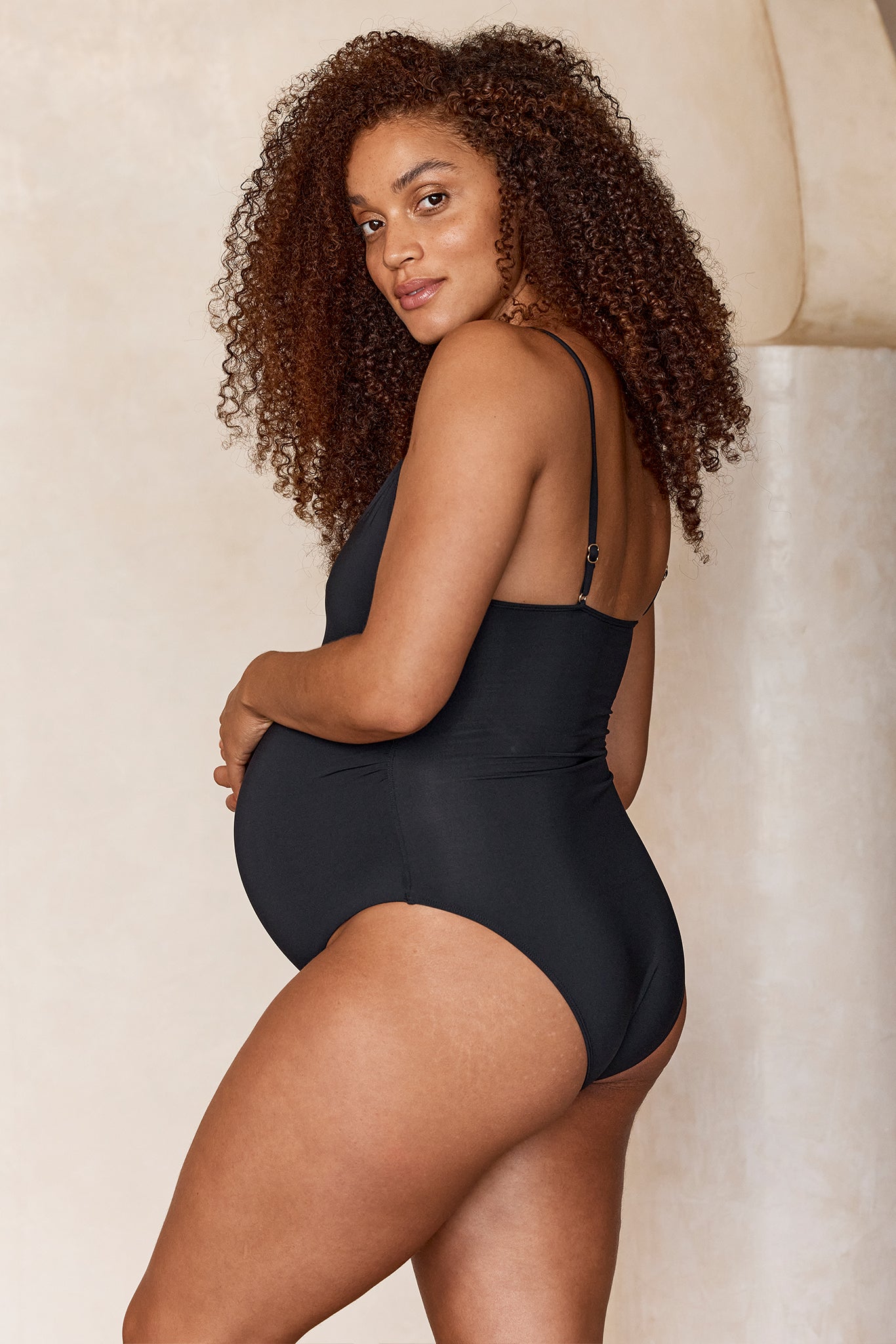 Pregnant woman in a black one-piece swimsuit, shown in profile with hands on belly, looking over her shoulder.