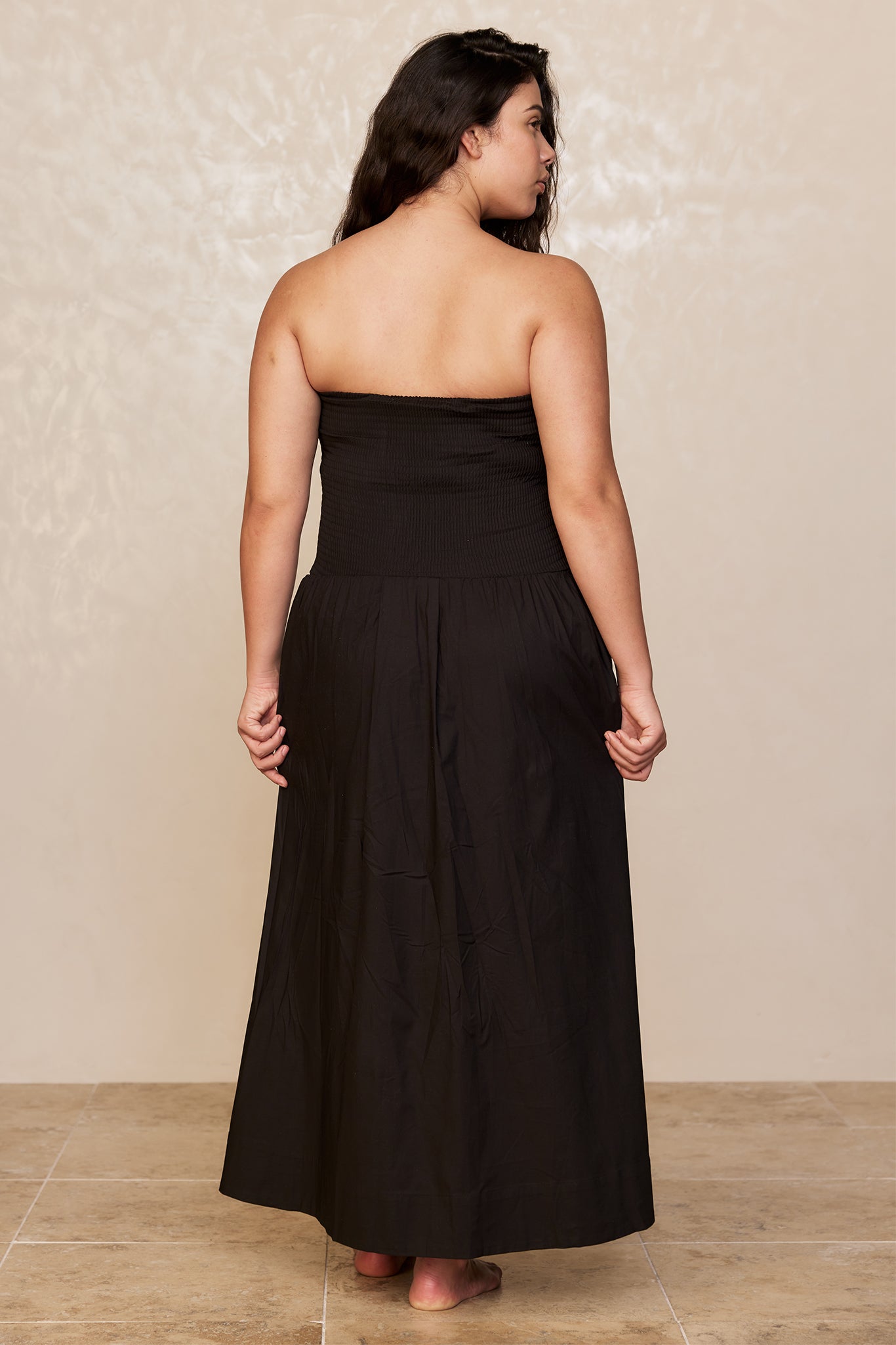 Back view of a woman wearing a strapless black maxi dress, barefoot on a tiled floor.