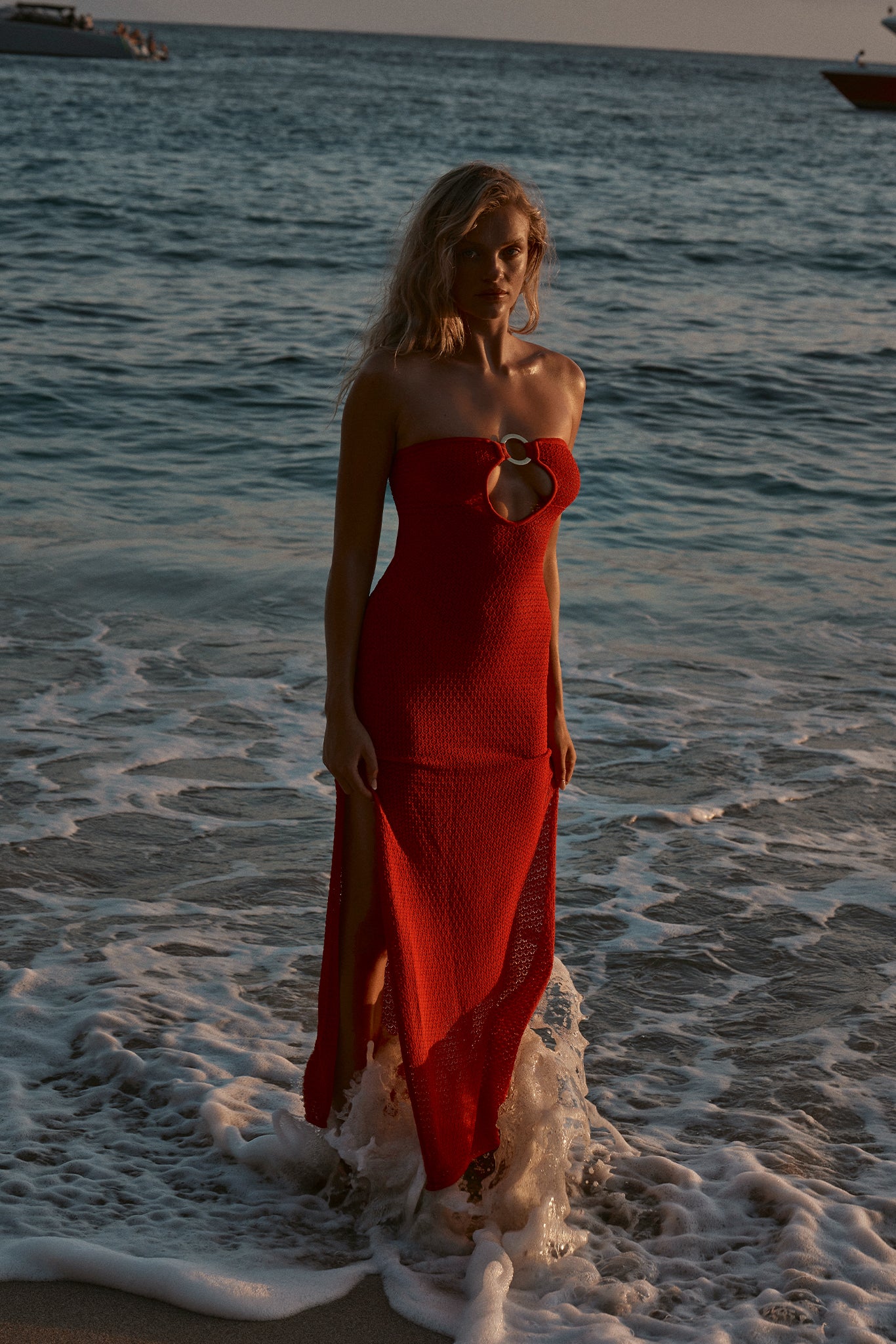 Woman standing knee-deep in ocean waves at sunset wearing a strapless red knit dress with a circular chest ring