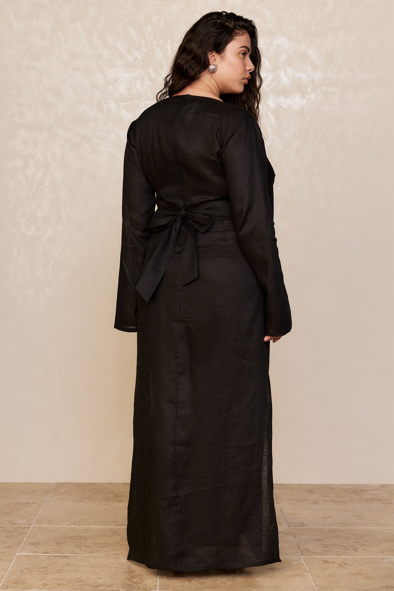 Back view of model wearing a long black linen dress with long sleeves and a tied waist bow, standing on tiled floor.
