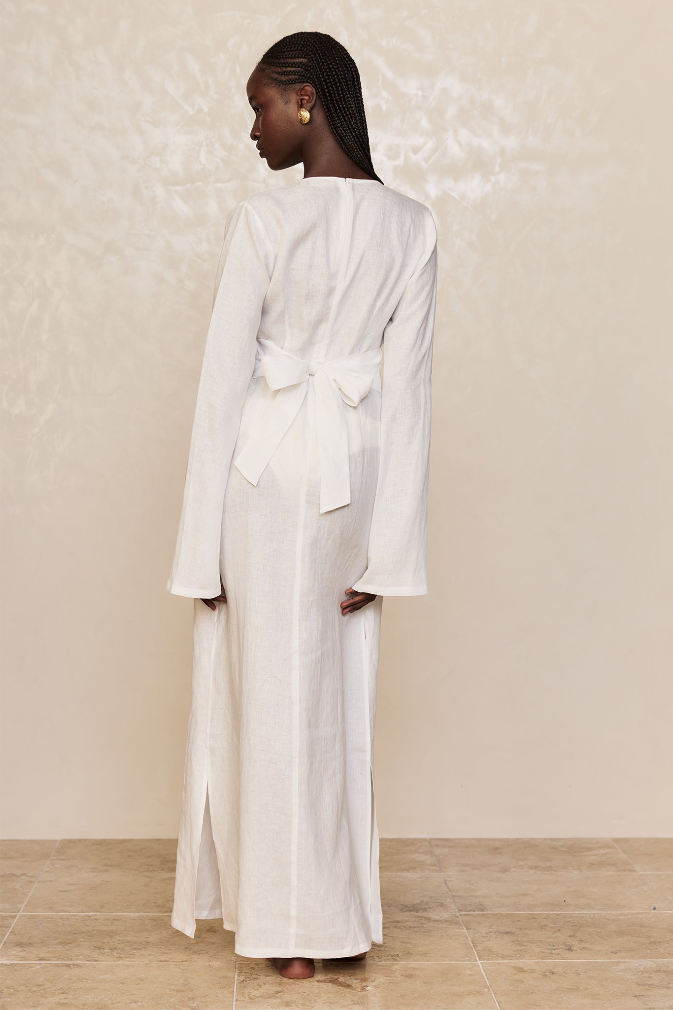 Model seen from back wearing long white linen dress with tied bow at lower back, braided hair and gold earrings.