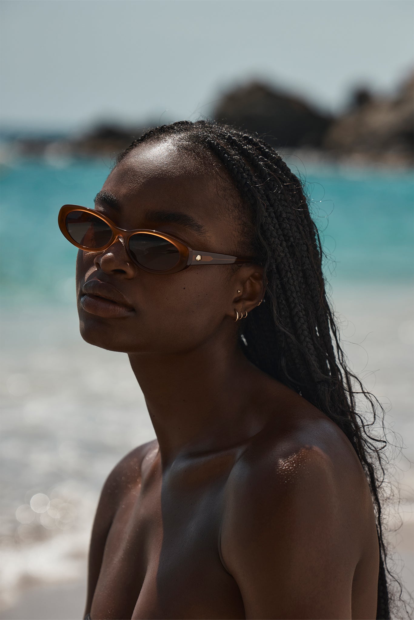 Black woman with braided hair wearing brown cat-eye sunglasses, sunlit beach in the background