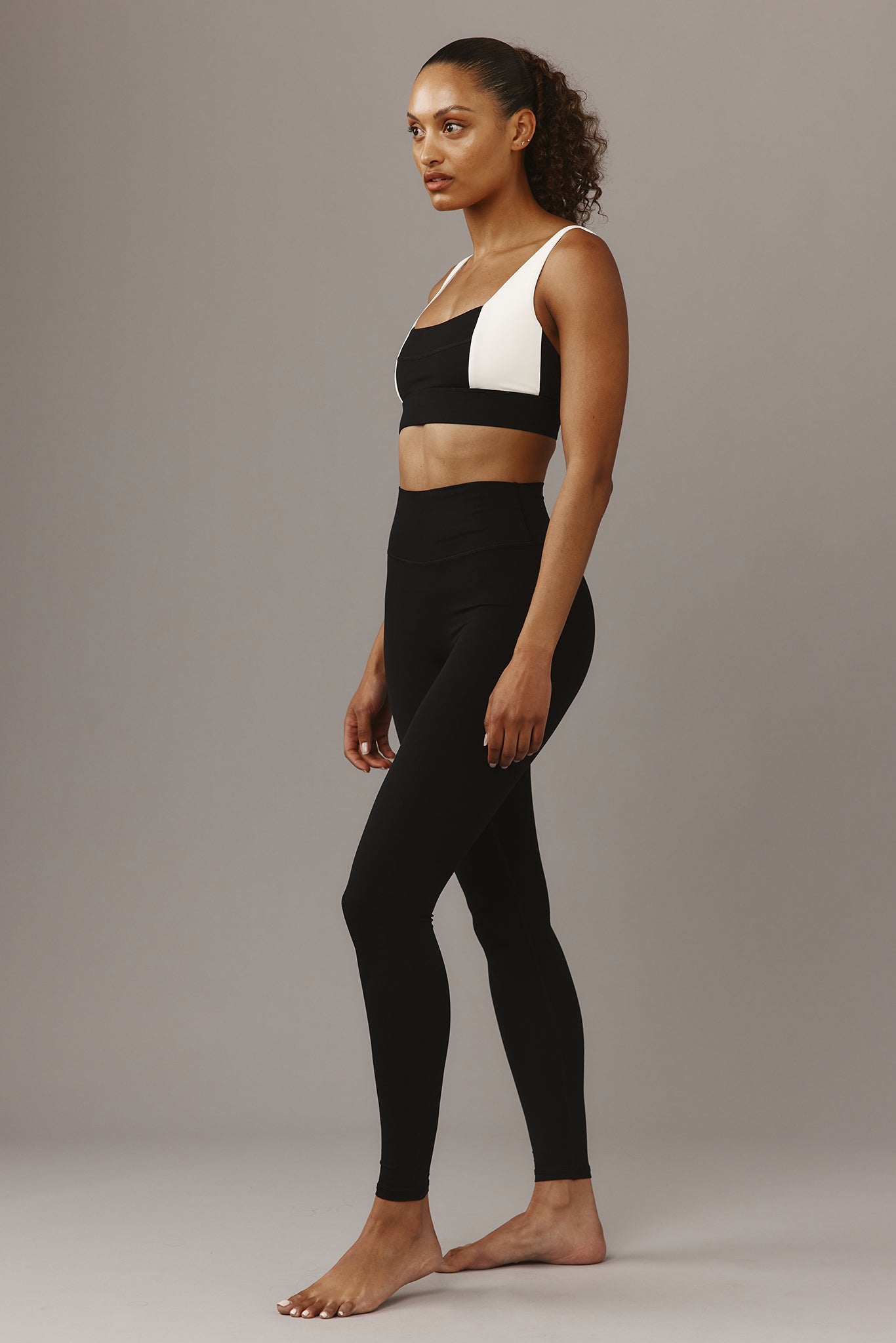 Female model in black high-waist leggings and black-and-white sports bra, standing in profile on neutral gray background