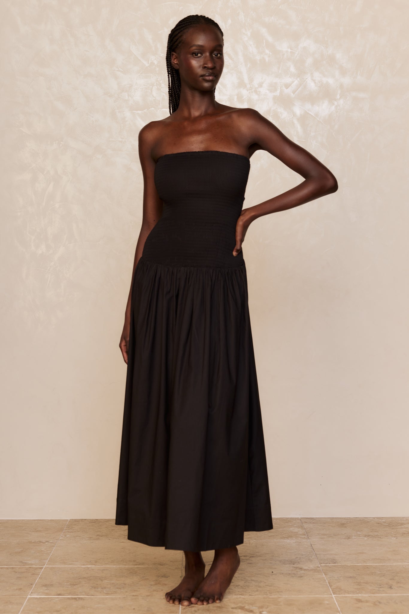 Model wearing strapless black maxi dress with smocked bodice and gathered skirt, standing barefoot against a cream backdrop.