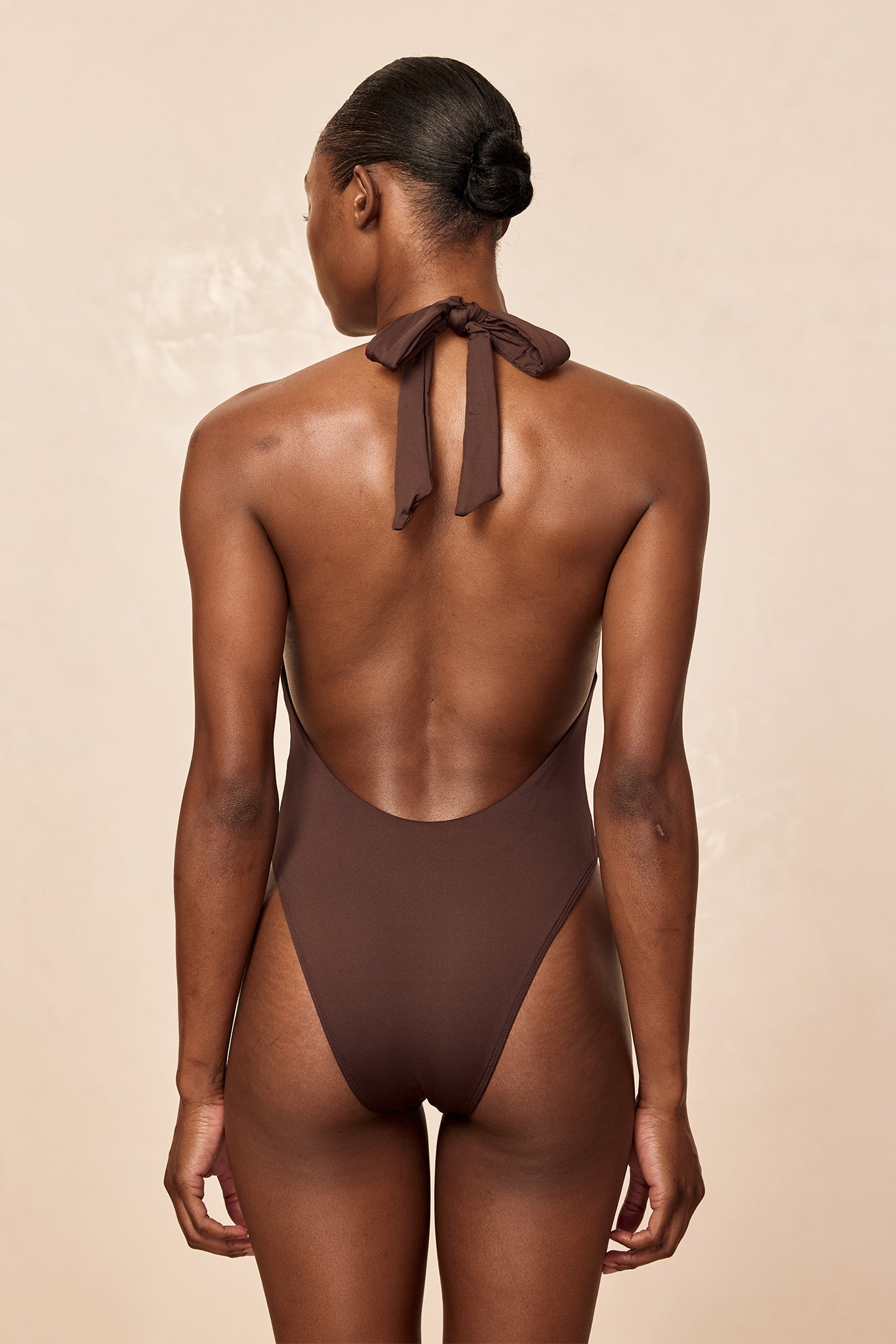 Back view of a model wearing a brown halter one-piece swimsuit tied at the nape, hair in a tight bun.