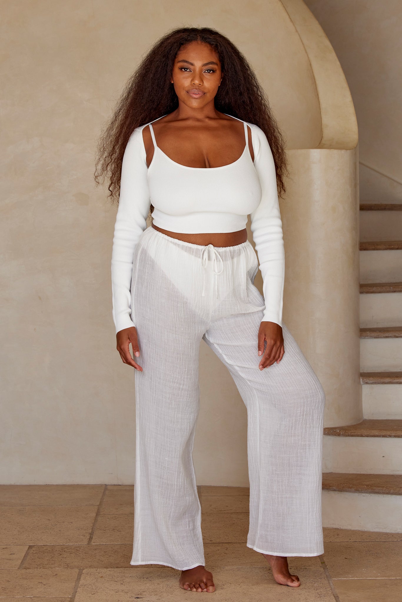Woman barefoot in white cropped top and sheer wide-leg drawstring pants by a curved staircase