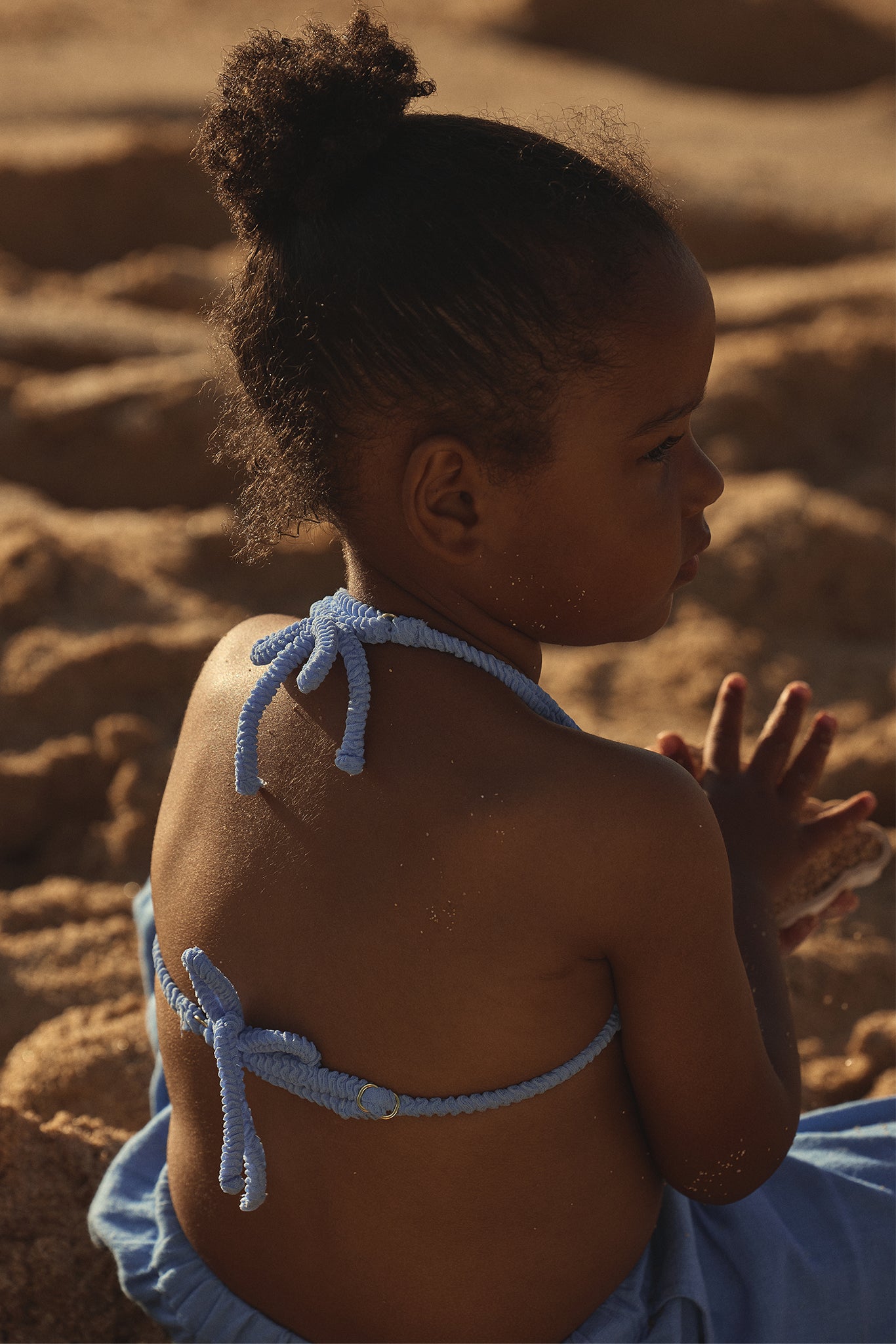 Young child sitting on a sandy beach seen from behind, wearing a light-blue halter bikini tied at neck and back, holding sand.