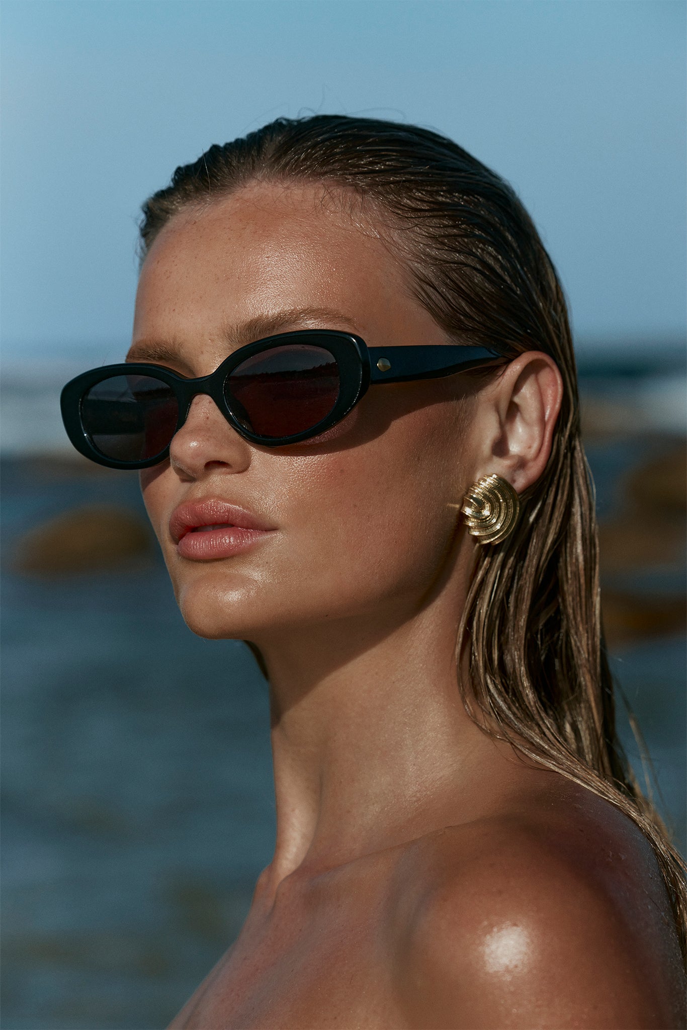 Woman portrait: black cat-eye sunglasses, gold spiral earring, wet hair, sunlit shoulder with blurred beach background.