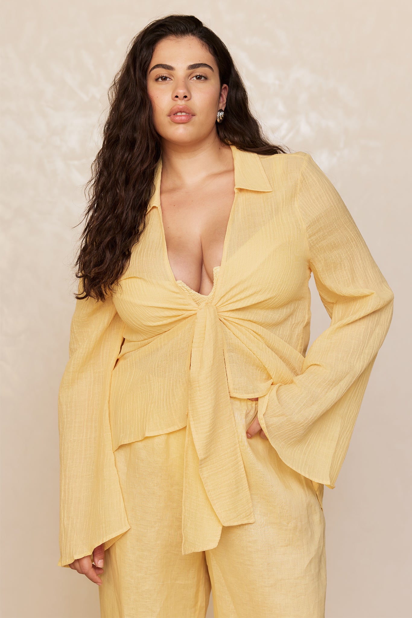 Plus-size woman with long wavy dark hair in a pale yellow tie-front blouse and matching pants, neutral background.