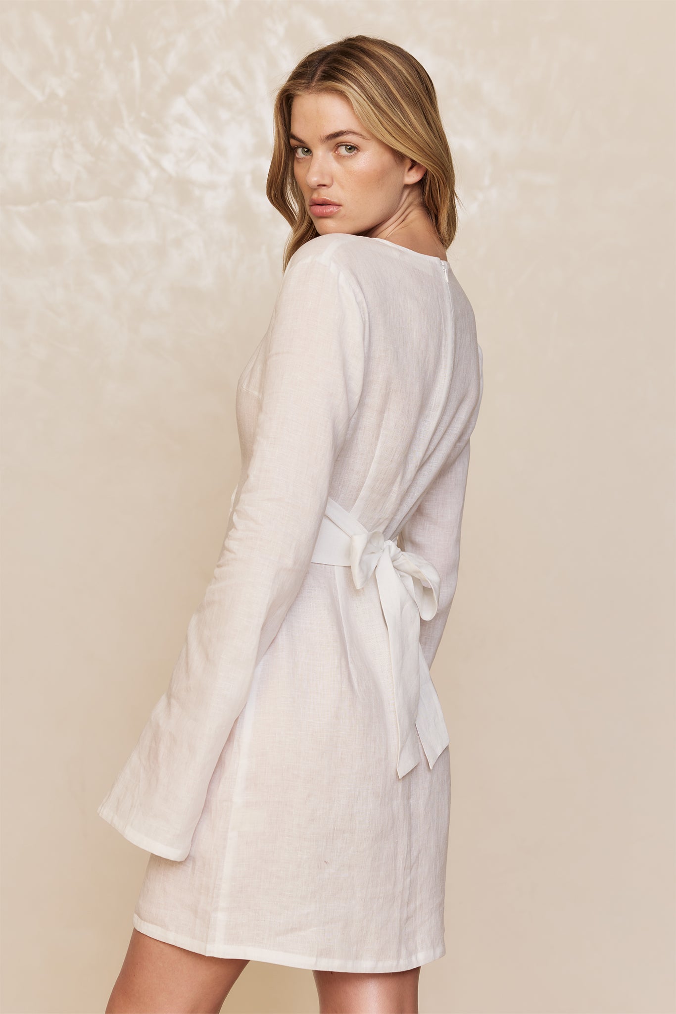 Back view of woman wearing a short white linen dress with long sleeves and a tied waist bow.