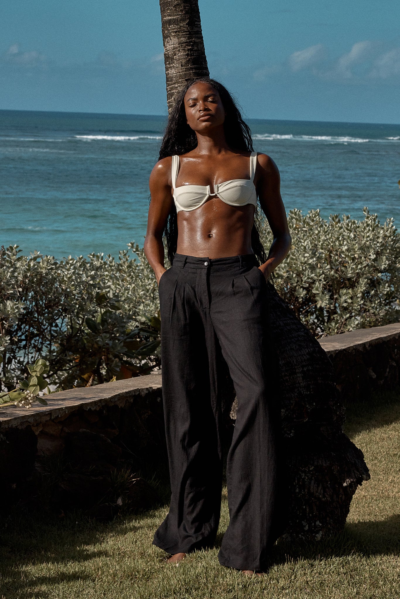 Black woman in a cream bikini top and black wide-leg pants leaning against a palm tree by the ocean, hands in pockets.