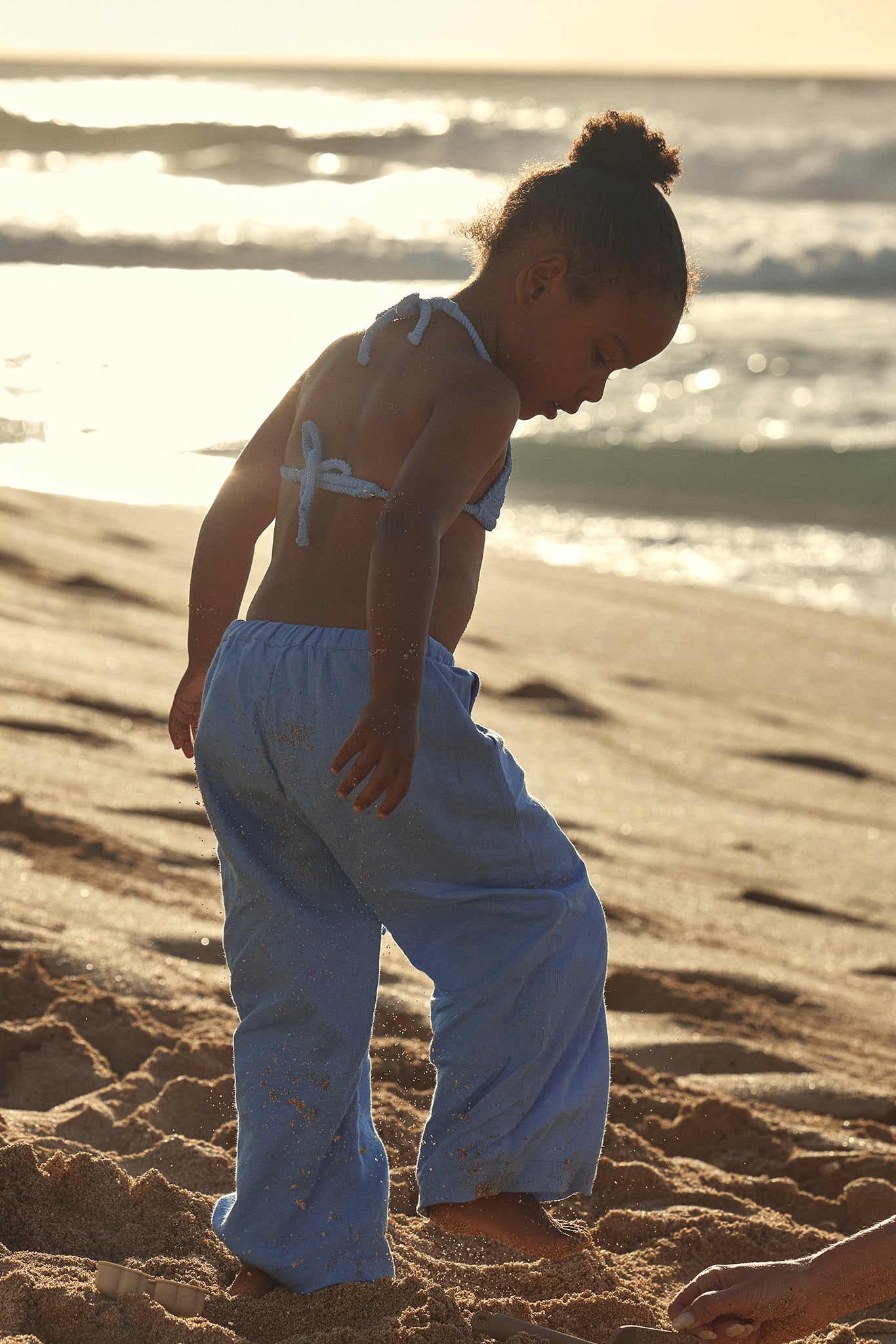 Child with hair in a bun wearing blue pants and a tied bikini top playing in sand on a sunlit ocean beach.