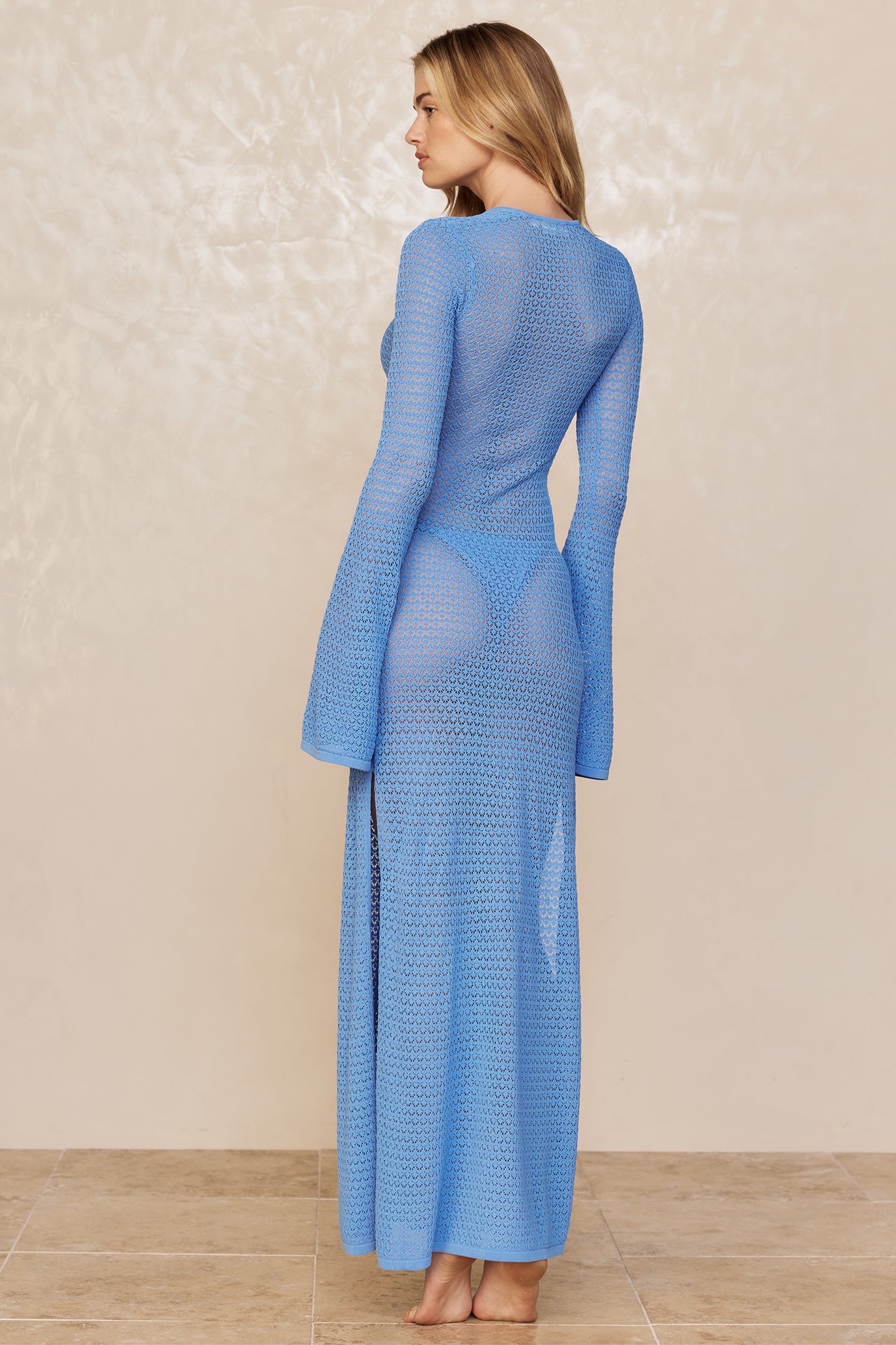 Woman standing barefoot on tile wearing a long sheer blue crochet dress, back and profile view.