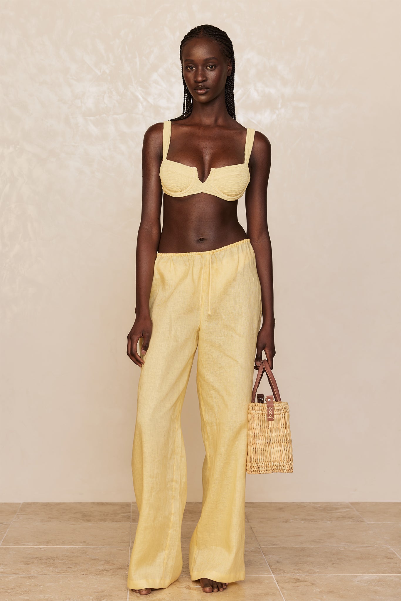 Model wearing yellow underwire bikini top and matching loose linen pants, holding a woven straw tote.