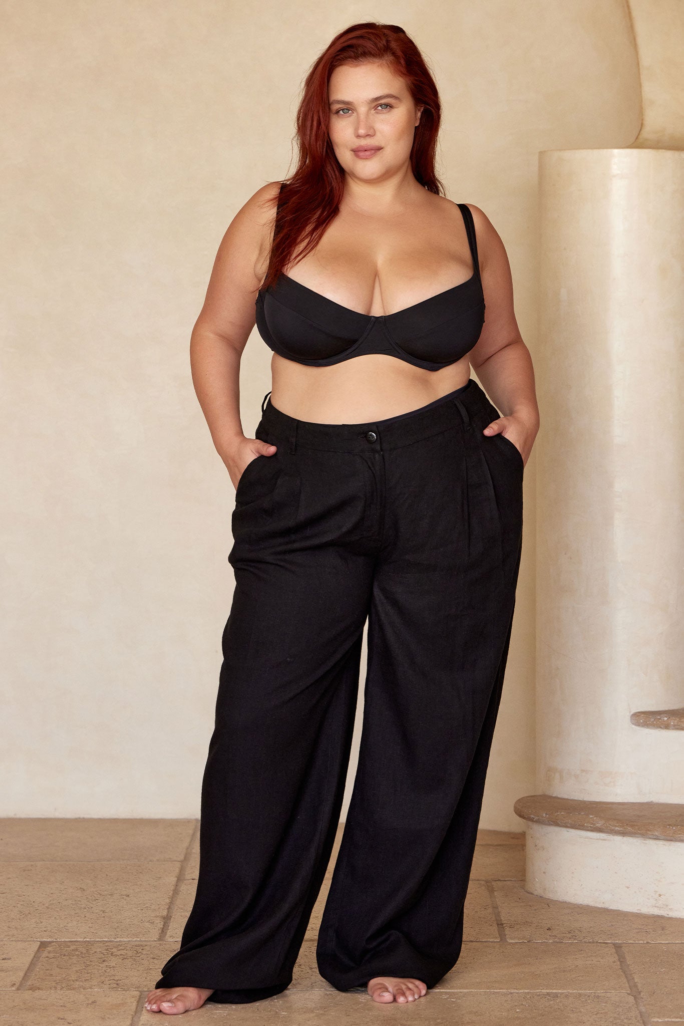 Plus-size model, long red hair, black underwire bra and wide-leg black pants, barefoot, hands in pockets.