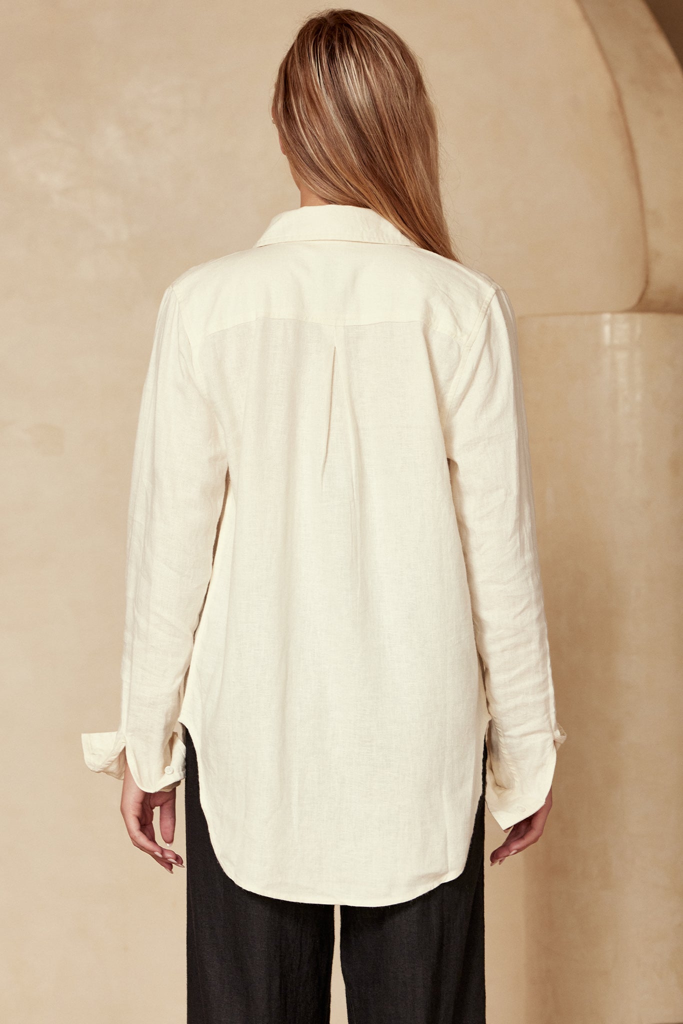 Back view of model wearing a cream long-sleeve button-up shirt with center back pleat, paired with black pants.