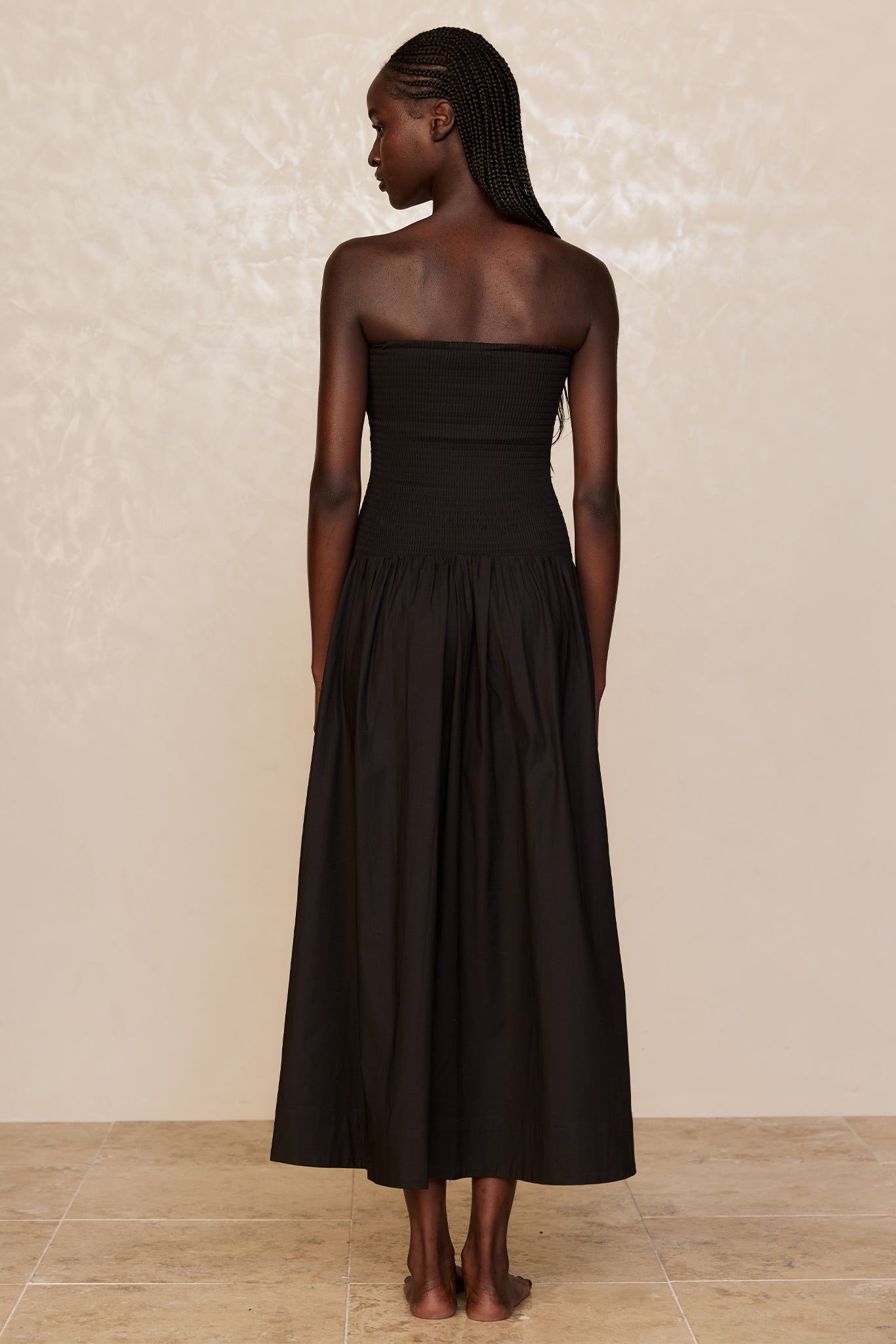 Back view of a woman with braids wearing a black strapless smocked bodice and long gathered skirt, barefoot on a tiled floor.