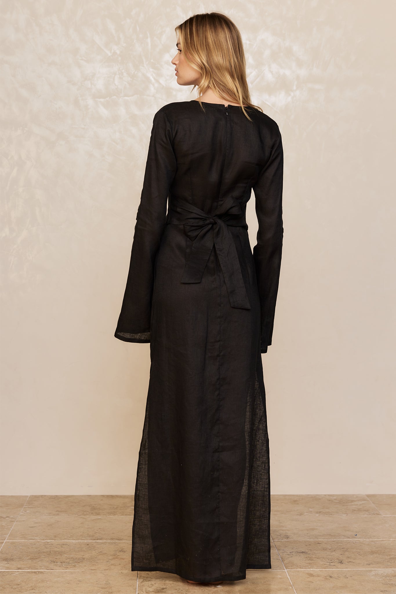 Back view of model wearing floor-length black linen dress with long sleeves and tied waist bow