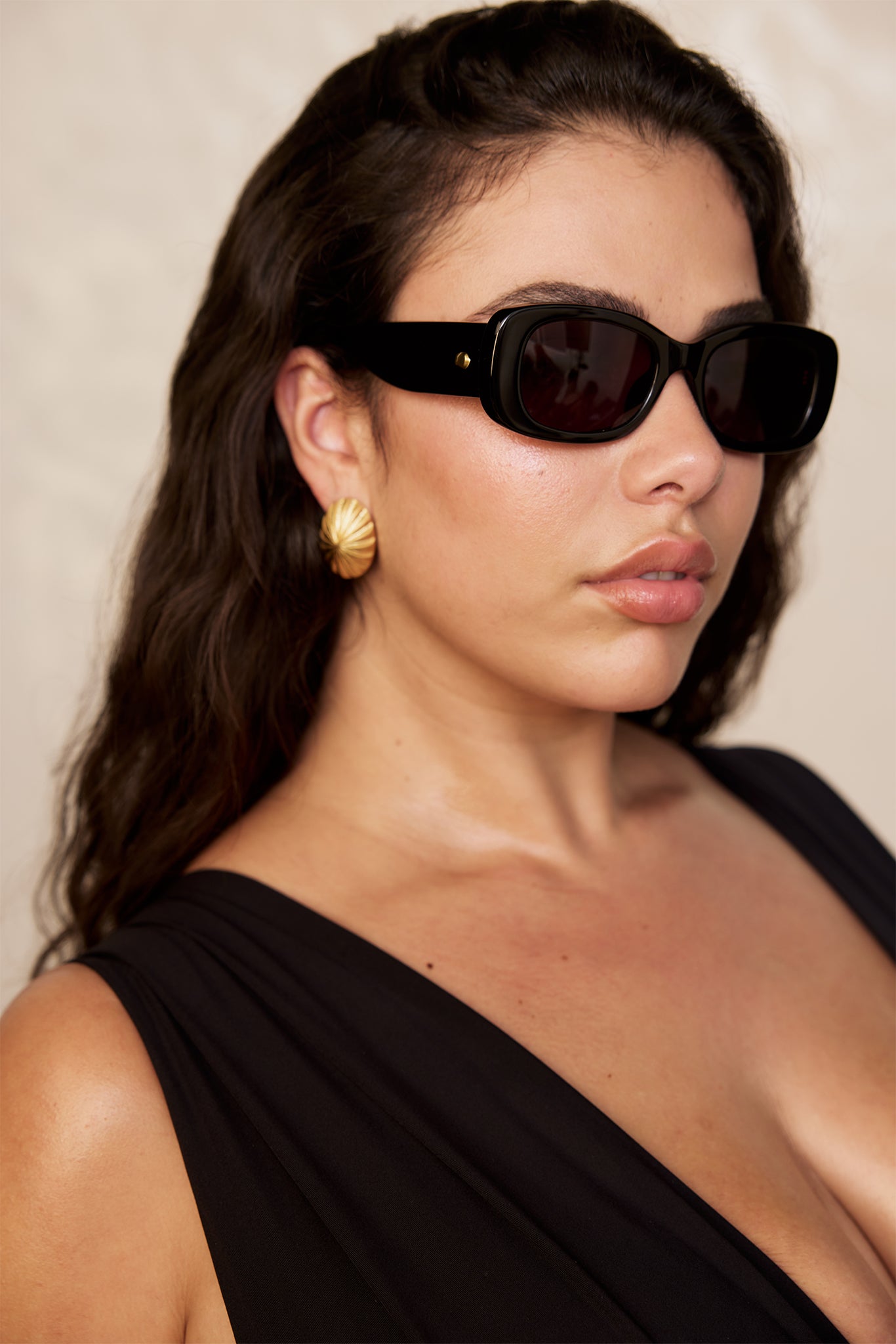 Person with dark hair wearing black rectangular sunglasses, gold shell earrings, black sleeveless top, shoulder-up portrait.