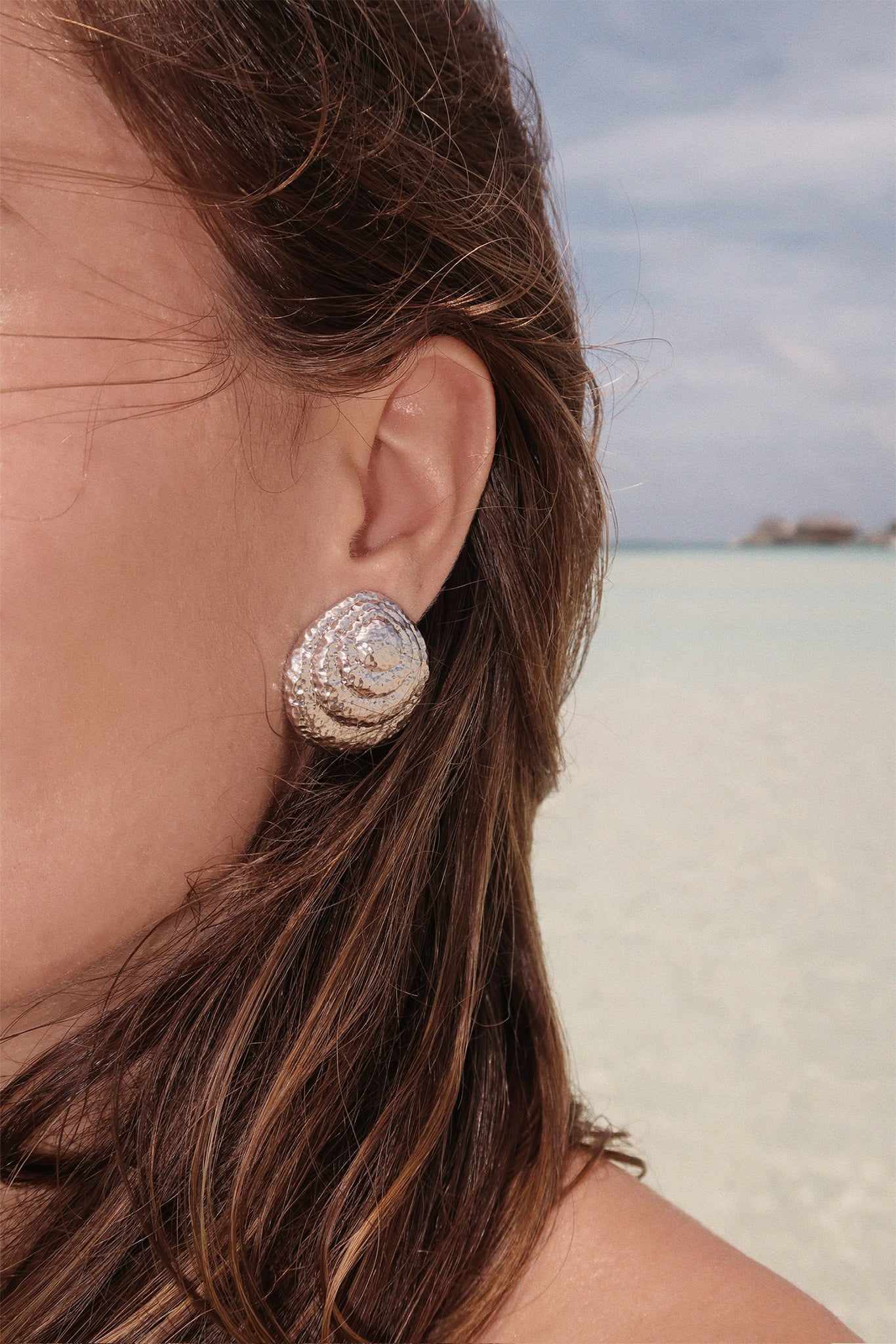 Close-up of woman's ear wearing a hammered spiral silver earring, with blurred beach and sea in the background.