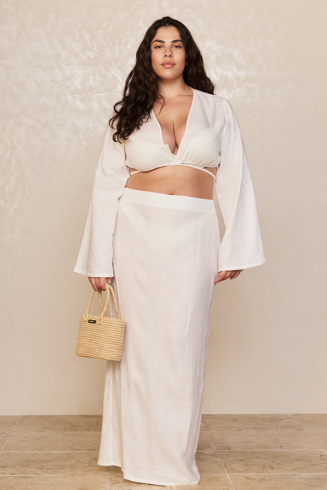 Plus-size model wearing a cream two-piece linen set and long skirt, holding a woven straw handbag against a textured beige backdrop.