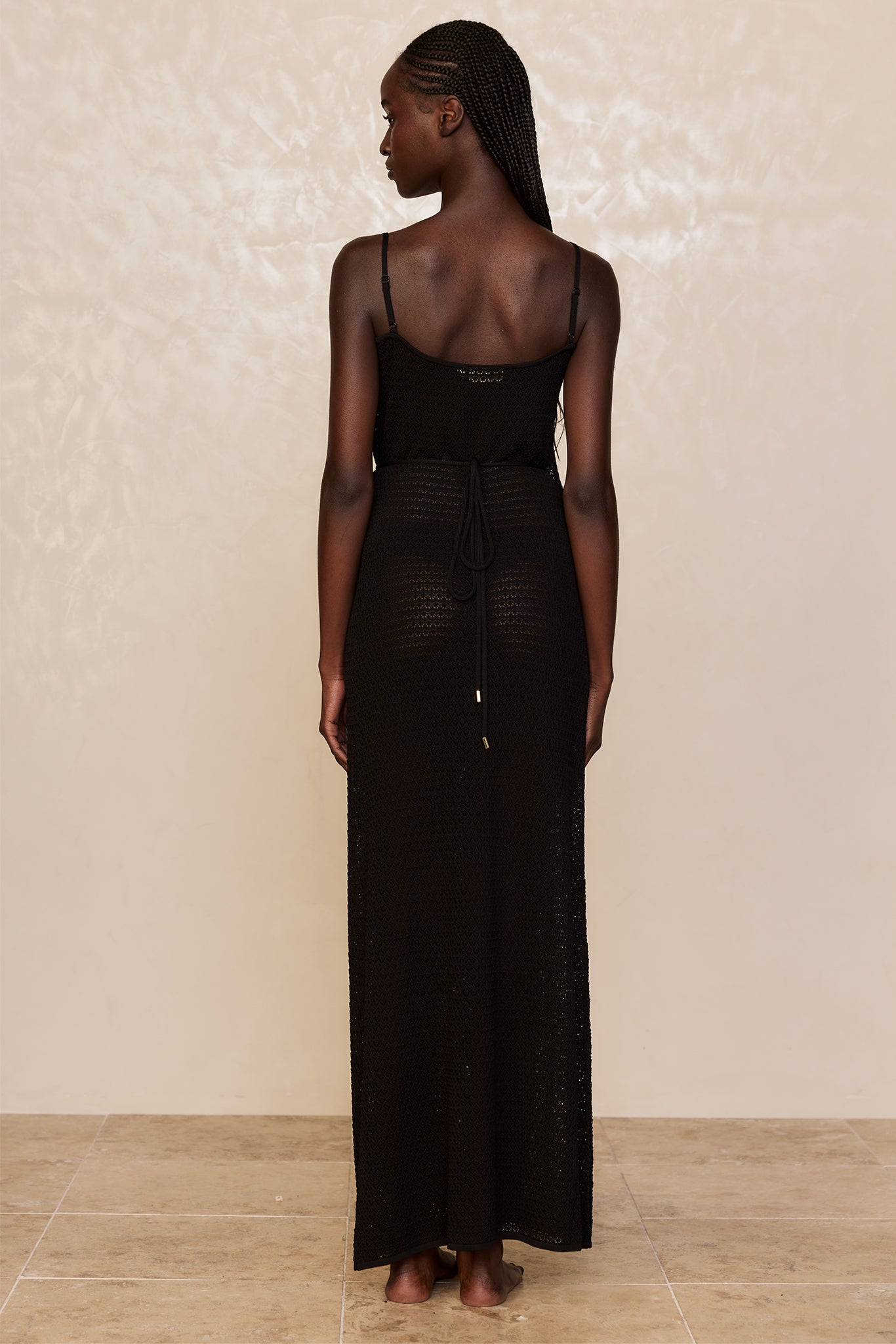 Back view of woman with long braids wearing a black crocheted maxi dress with waist tie, standing barefoot on tiled floor.