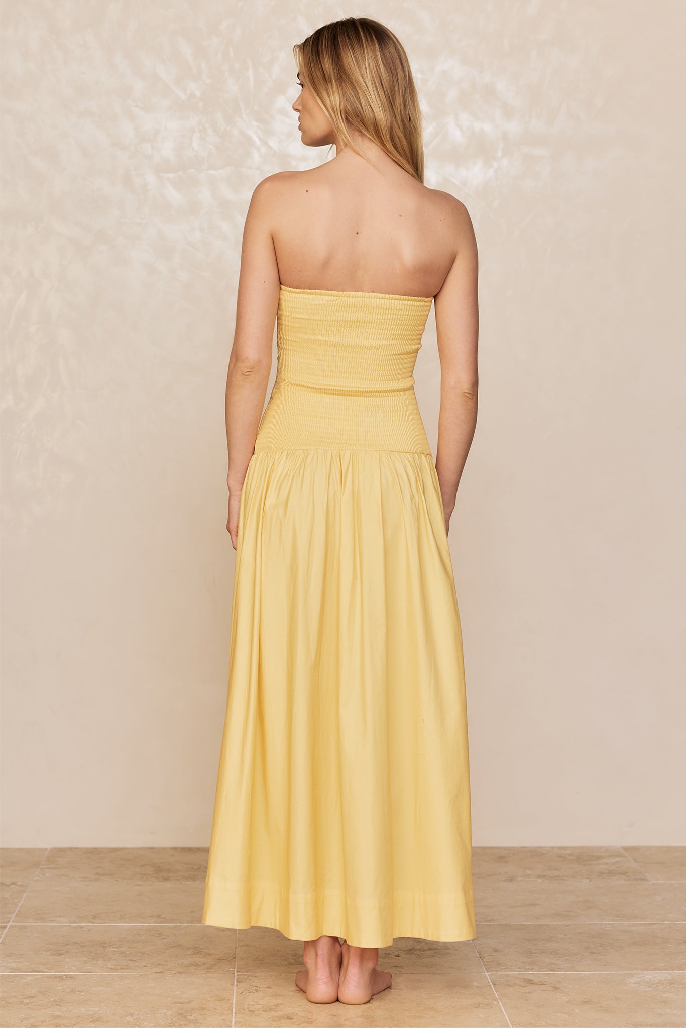 Woman standing barefoot, back view, wearing strapless yellow maxi dress on beige backdrop and tile floor.