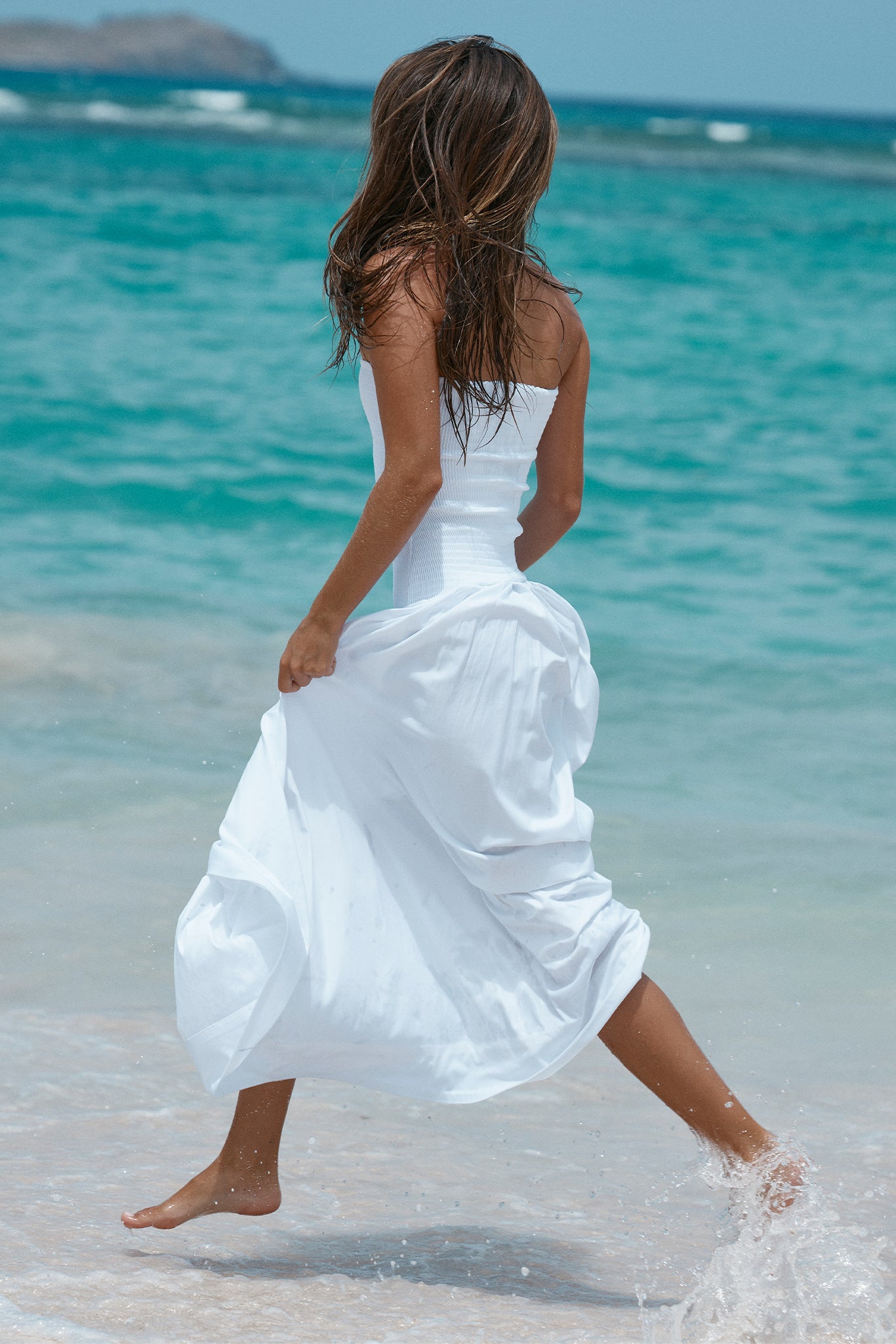 Woman in a white strapless dress running along a turquoise ocean shoreline, bare feet splashing in shallow water, back turned.
