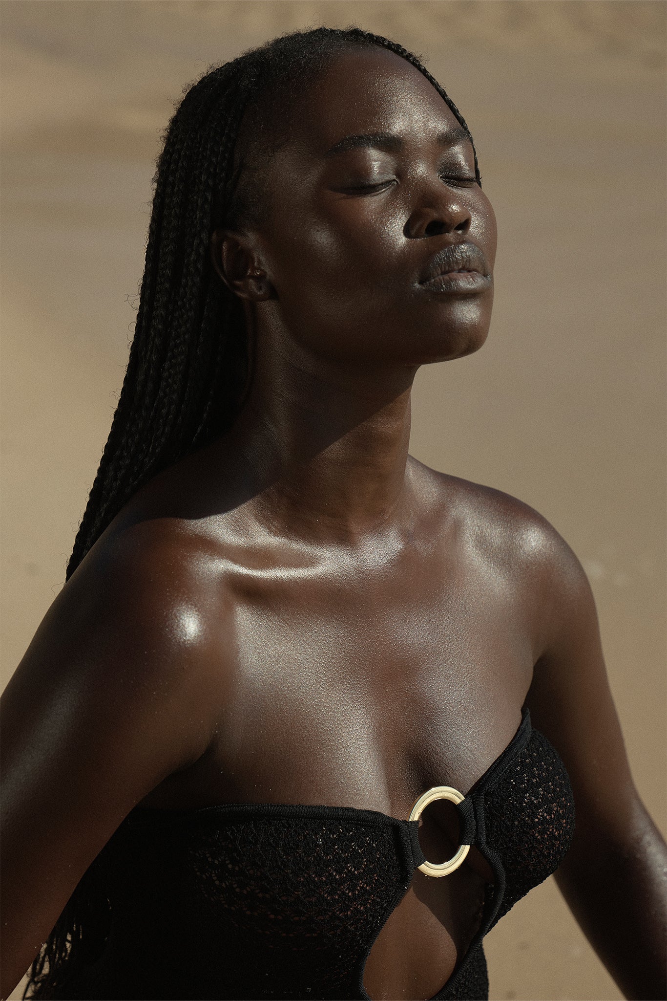 Black woman with long braids, eyes closed, sunlit dewy skin, wearing a black strapless swimsuit with central gold ring
