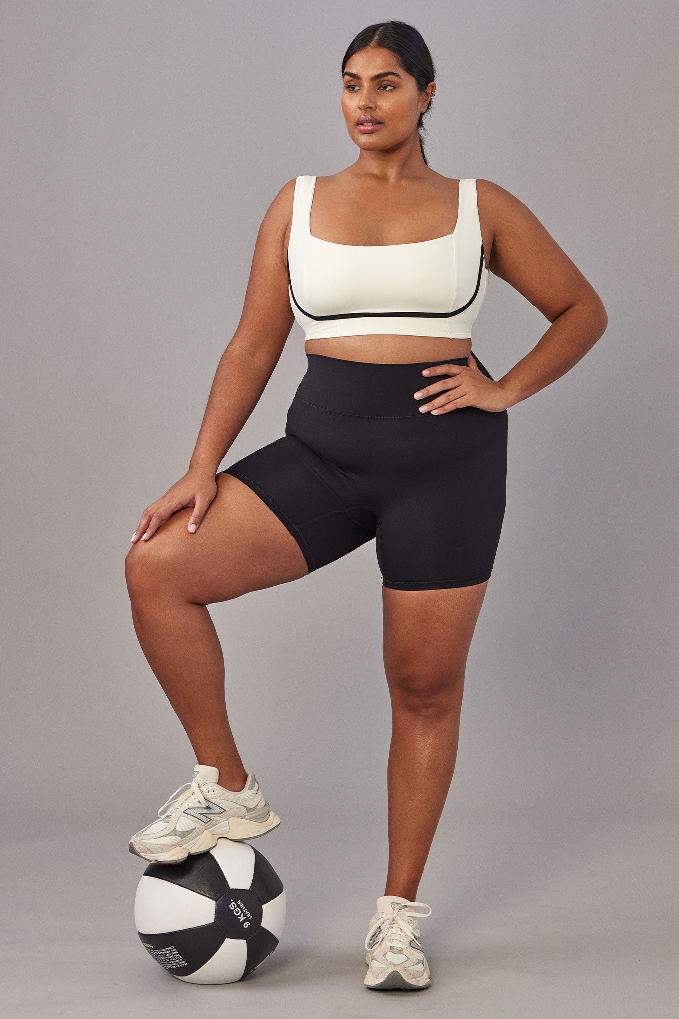 Plus-size model in cream sports bra and black high-waist bike shorts, left foot on black-and-white ball labeled 9 KGS.