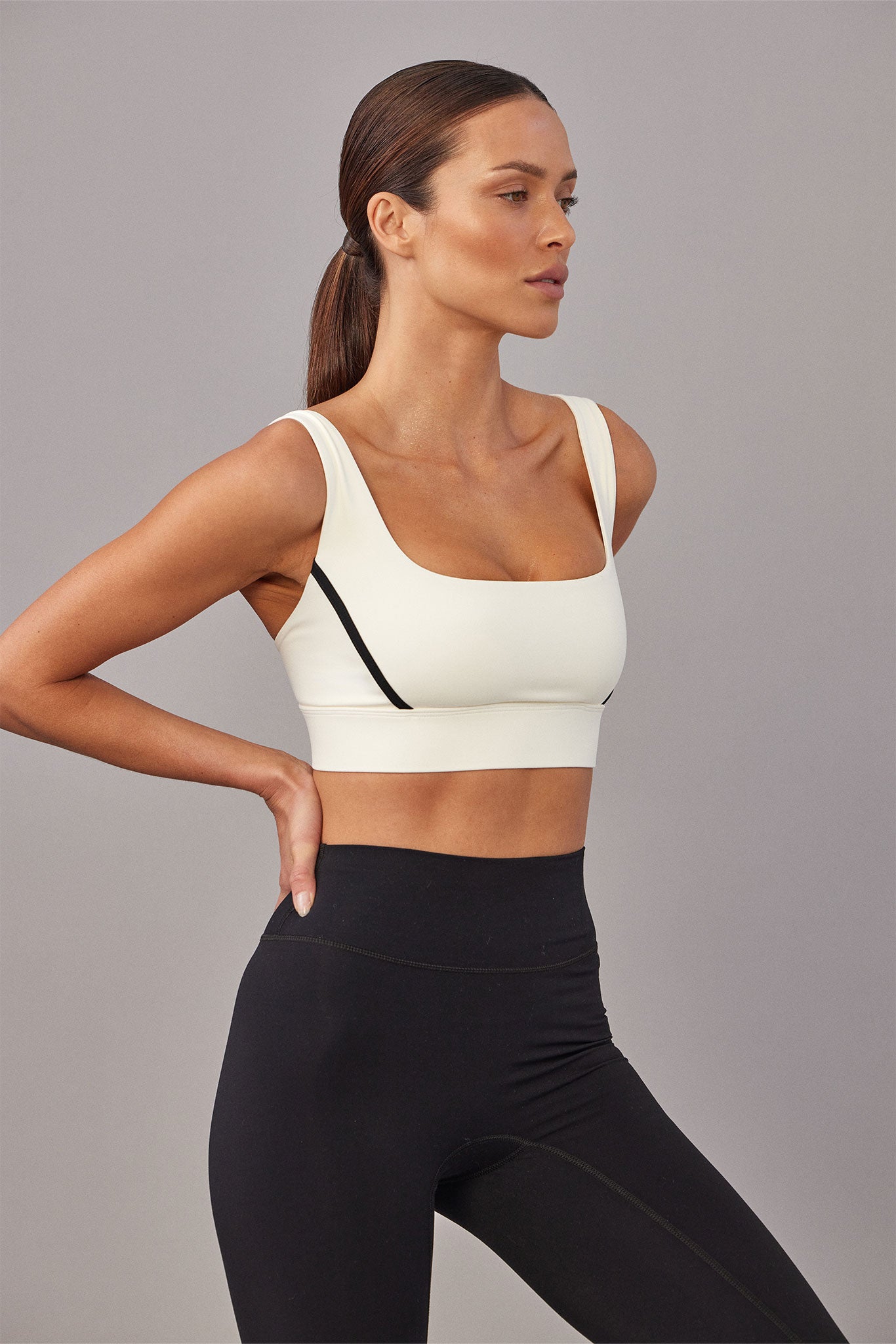 Side-profile woman wearing cream sports bra with black trim and high-waisted black leggings, hand on hip, gray backdrop.