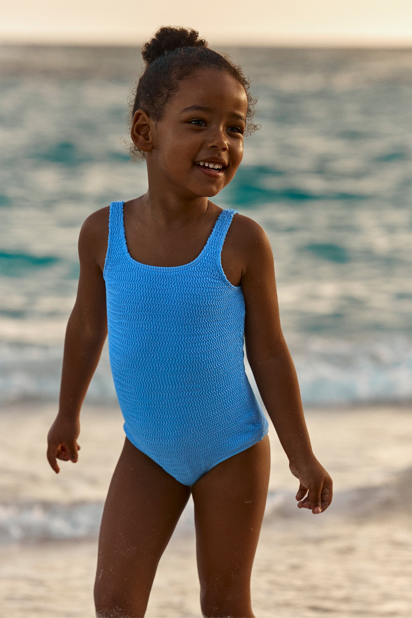 Smiling young girl in a textured sky-blue one-piece swimsuit standing on a sandy beach with waves in the background.