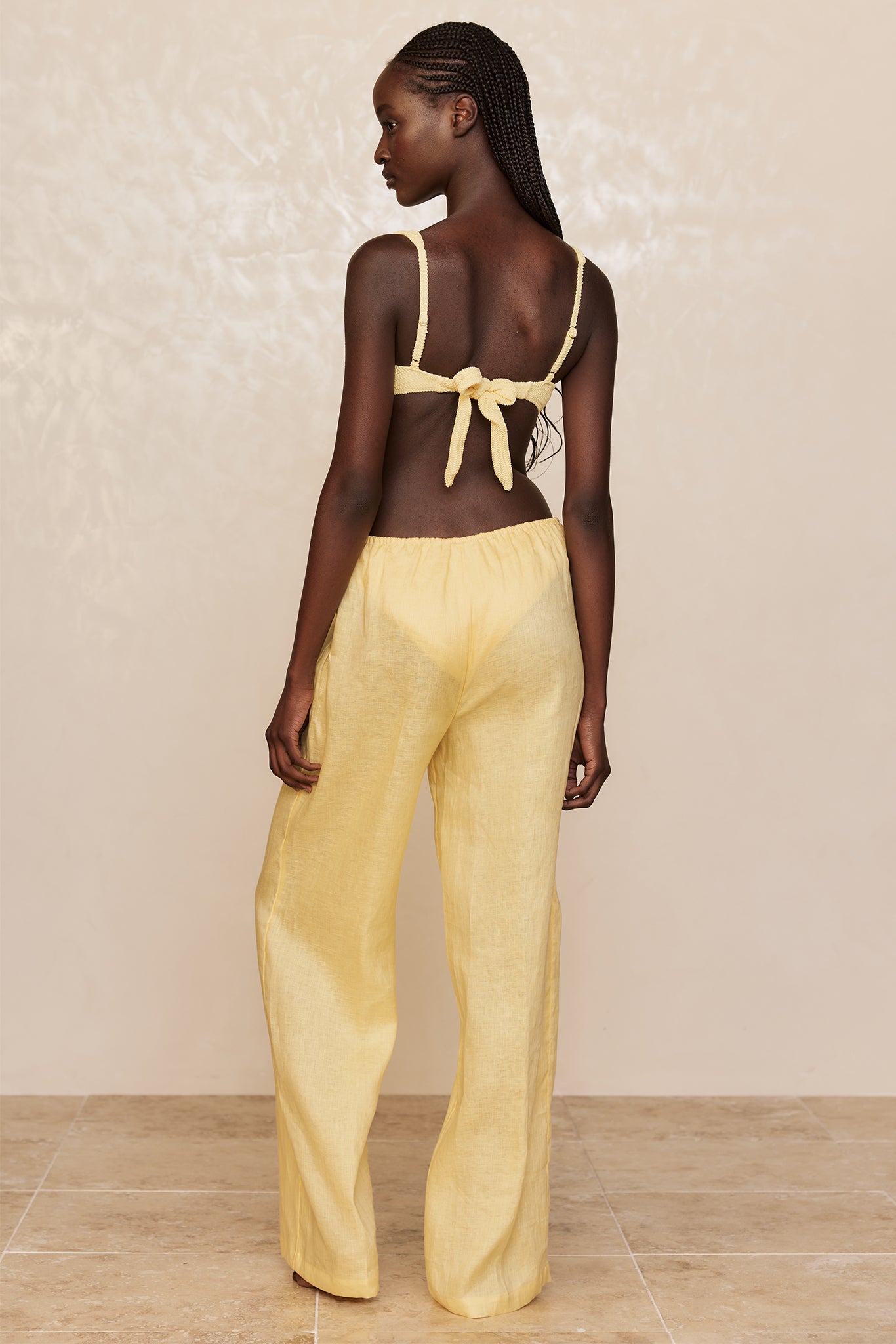 Model back view wearing yellow tie-back bikini top and loose yellow linen trousers on beige backdrop and tile floor.