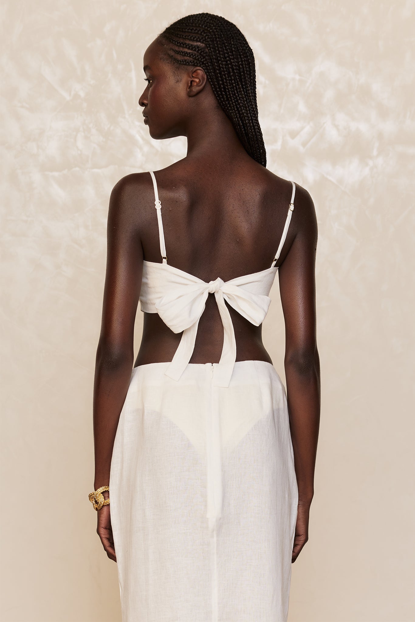 Rear view of model with cornrow braids wearing a white tie-back crop top and semi-sheer white skirt with a gold bracelet