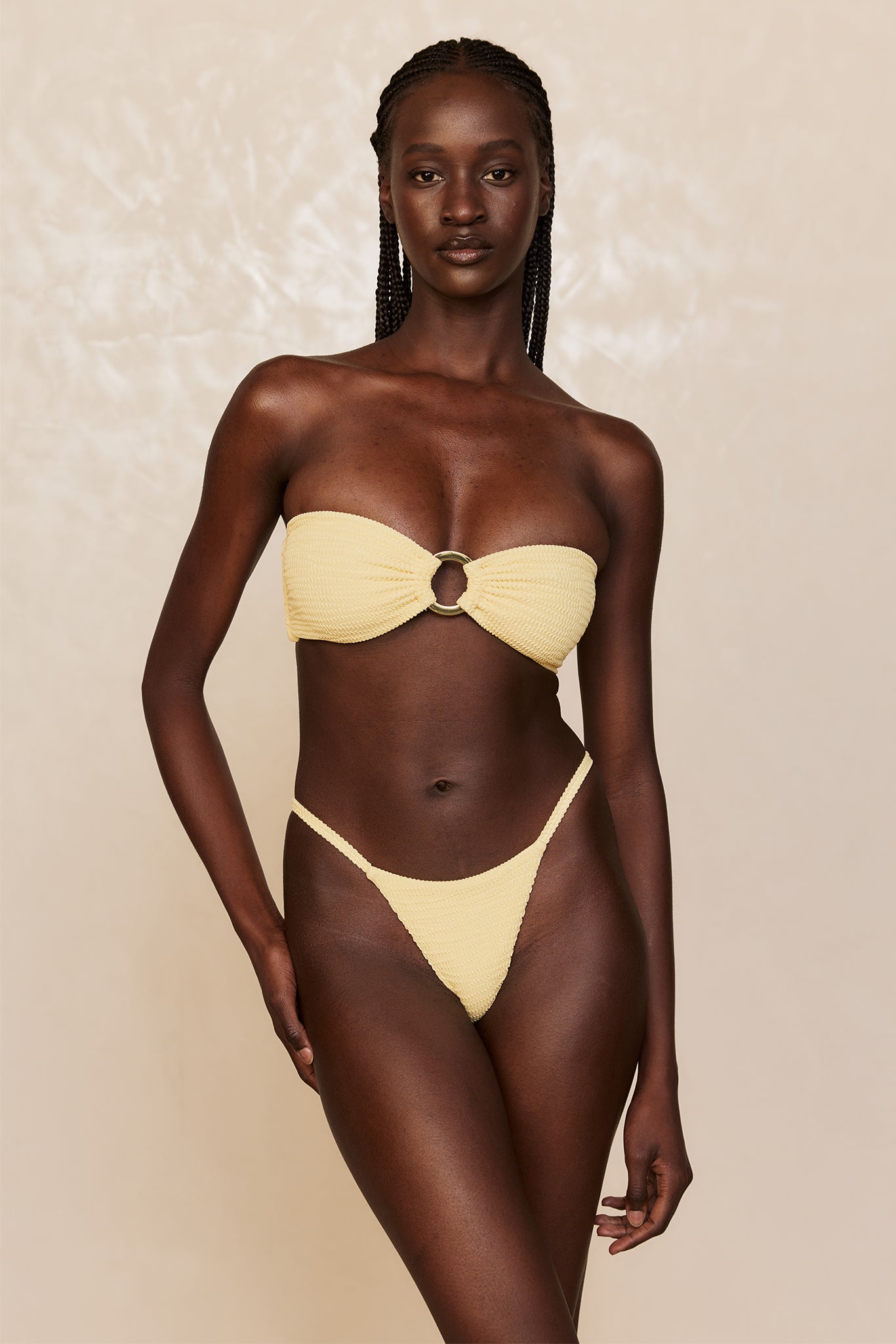 Model wearing pale-yellow textured bandeau bikini with center ring and high-cut bottoms on neutral background.