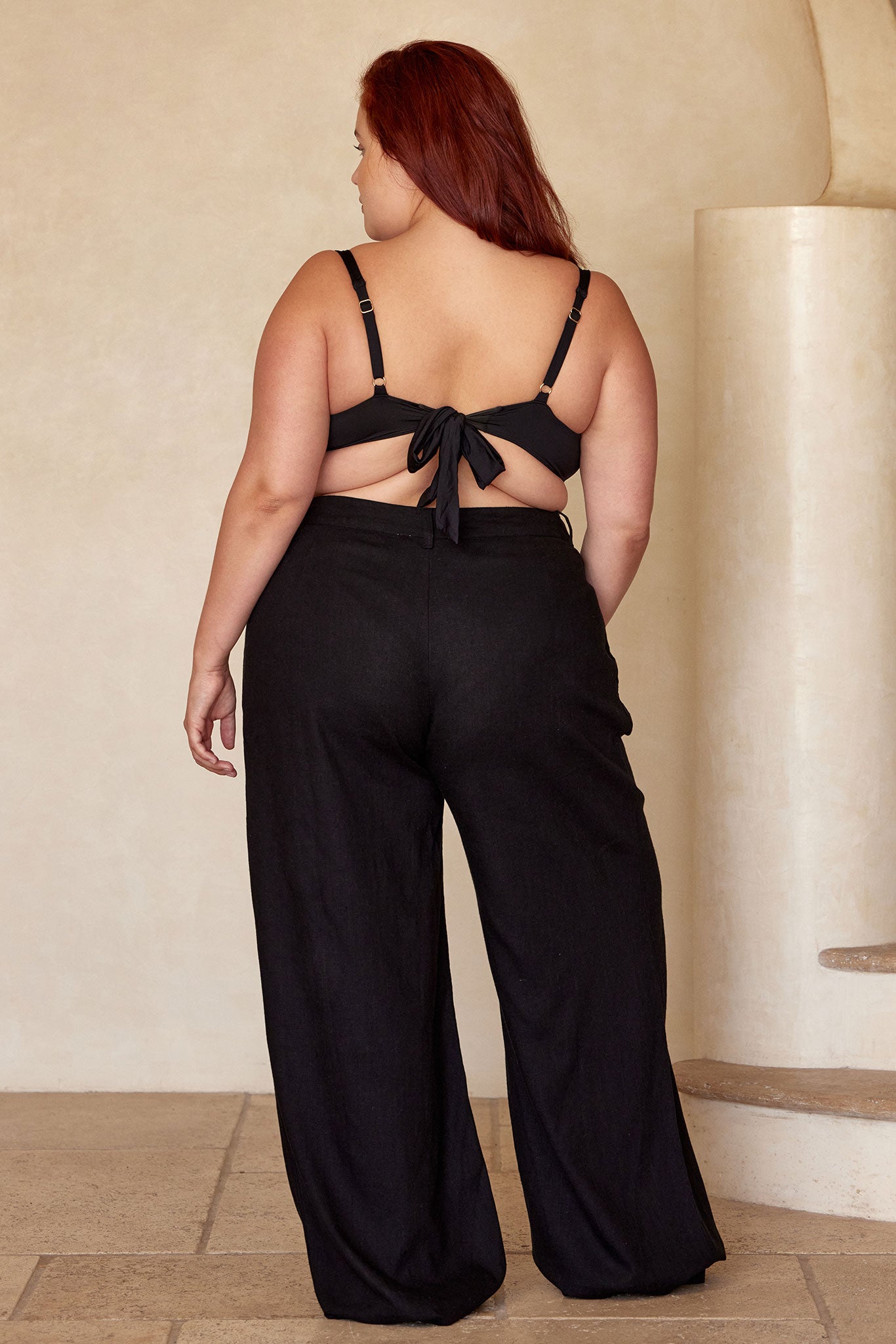 Plus-size woman seen from back wearing black tie-back bikini top and high-waisted wide-leg black pants against a beige plaster wall.