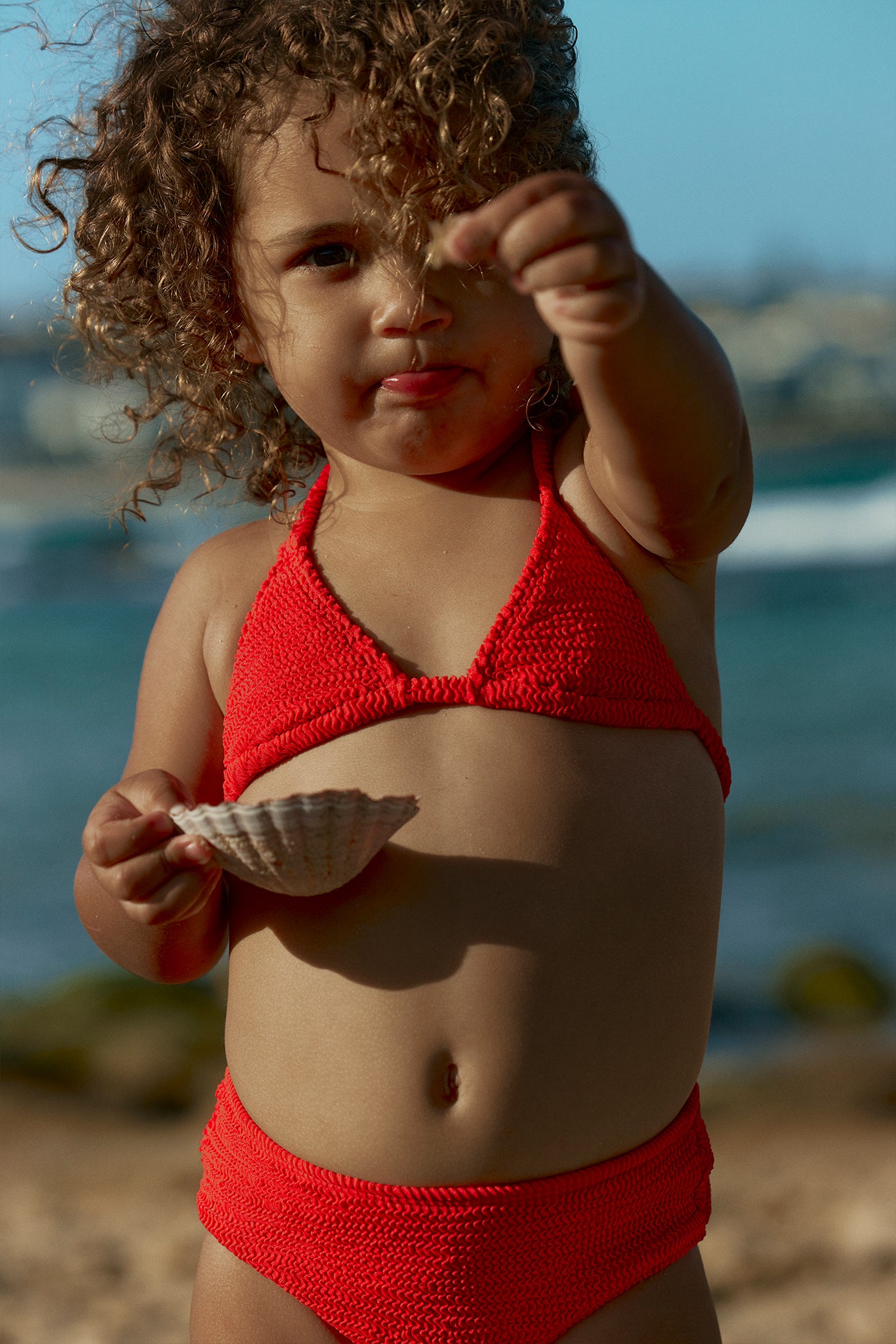 Toddler with curly hair at the beach holding a scallop shell, wearing a red knit bikini.
