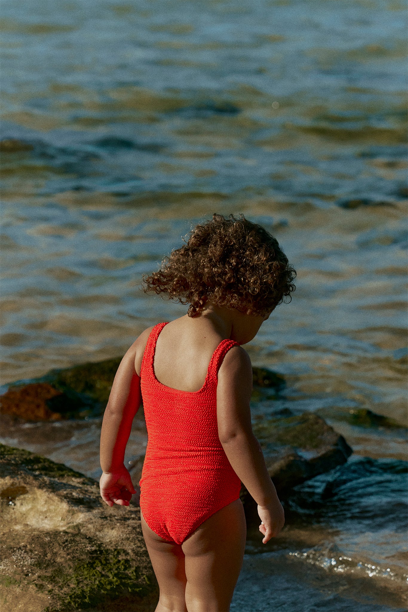 Young child in a red one-piece swimsuit standing on rocky shoreline, facing the water.